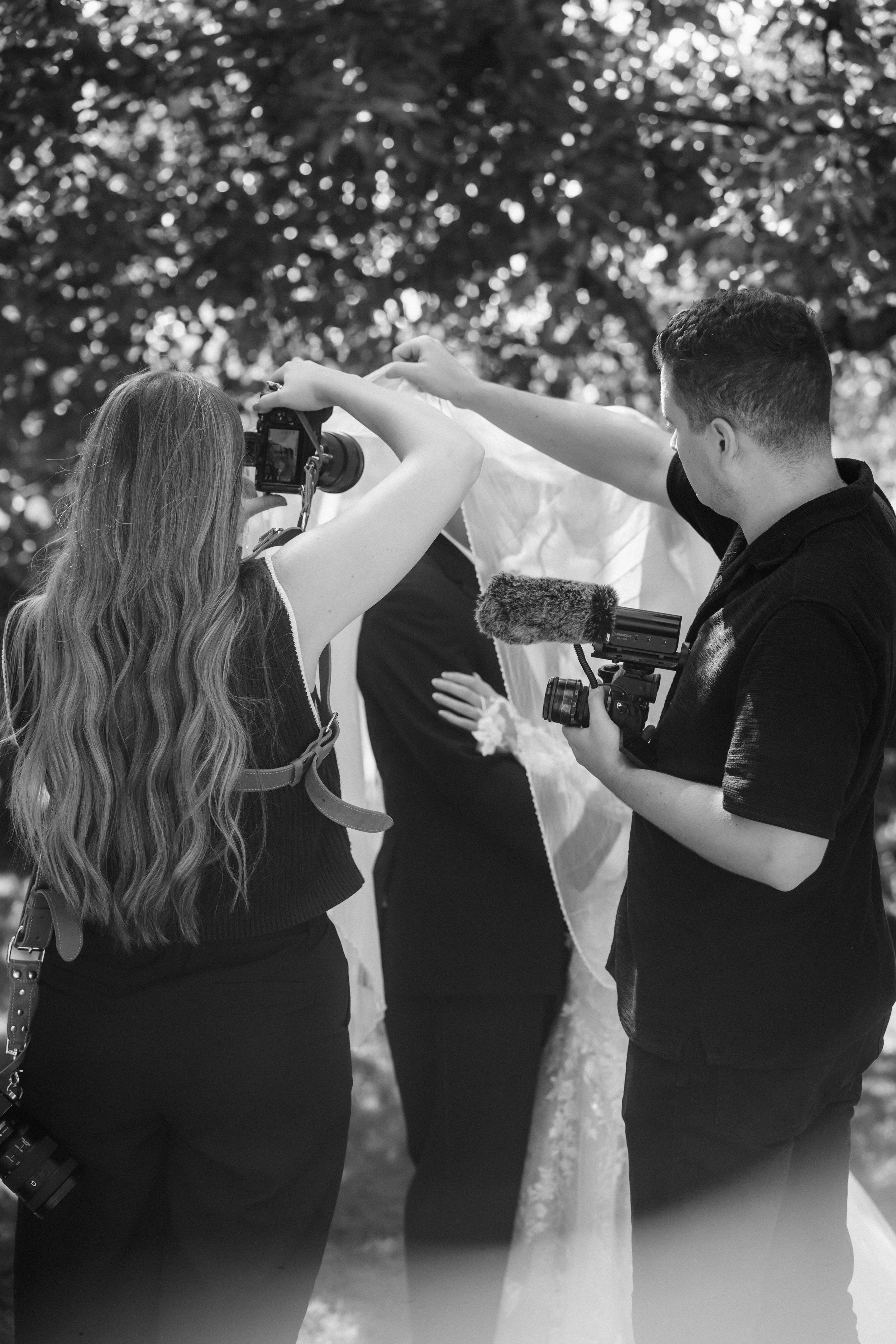 A bride and groom holding hands in an outdoor setting with trees, the bride in a white gown holding a bouquet and the groom in a suit with a bow tie.