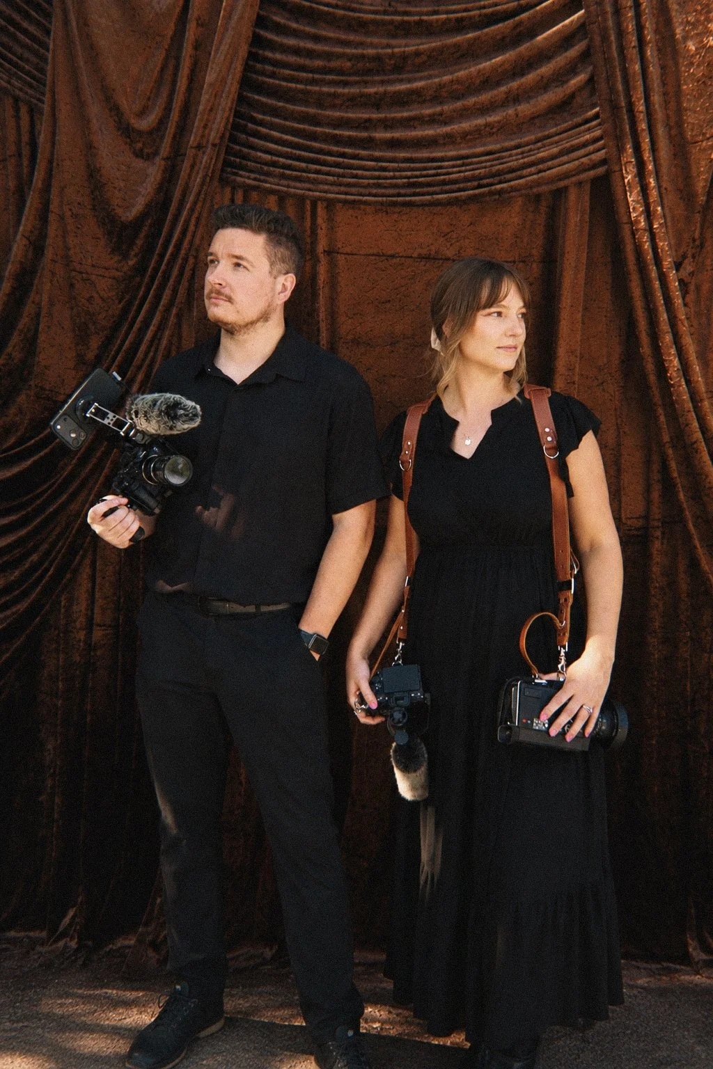 A man and a woman standing in front of a brown curtain, both holding cameras, with the man holding a DSLR camera with a microphone, and the woman holding a vintage camera with a strap, both dressed in black.