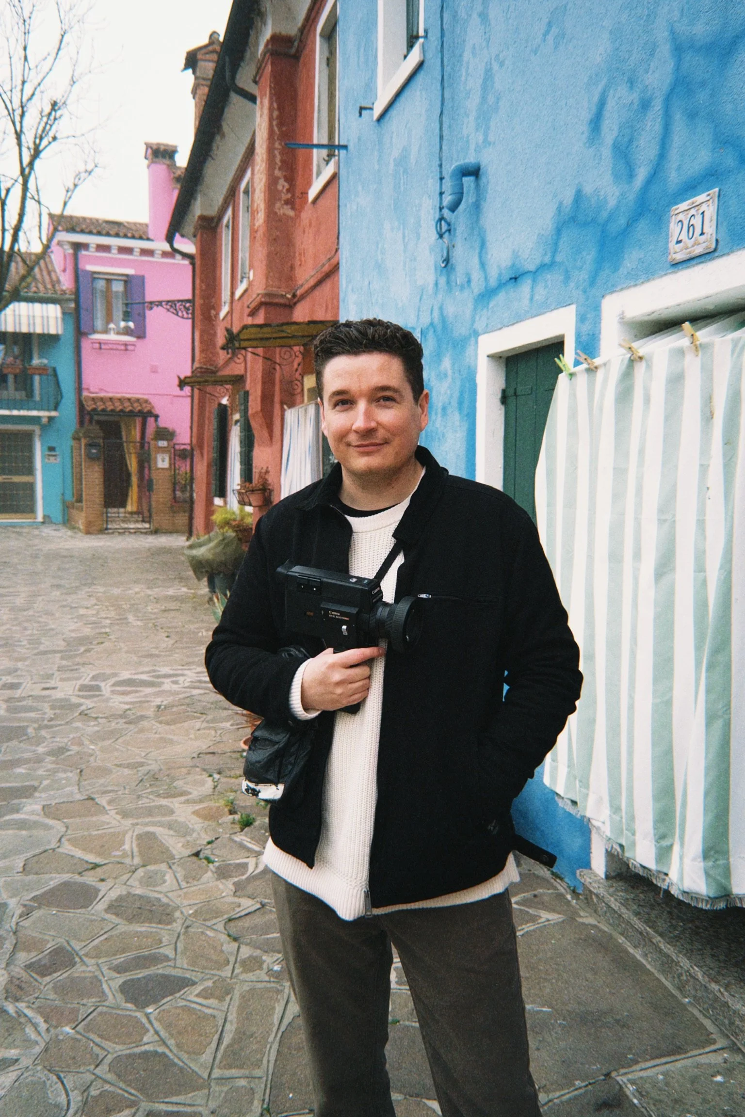 A person wearing a white shirt and black pants standing outdoors in a rocky, natural setting with dry grass and shrubbery.