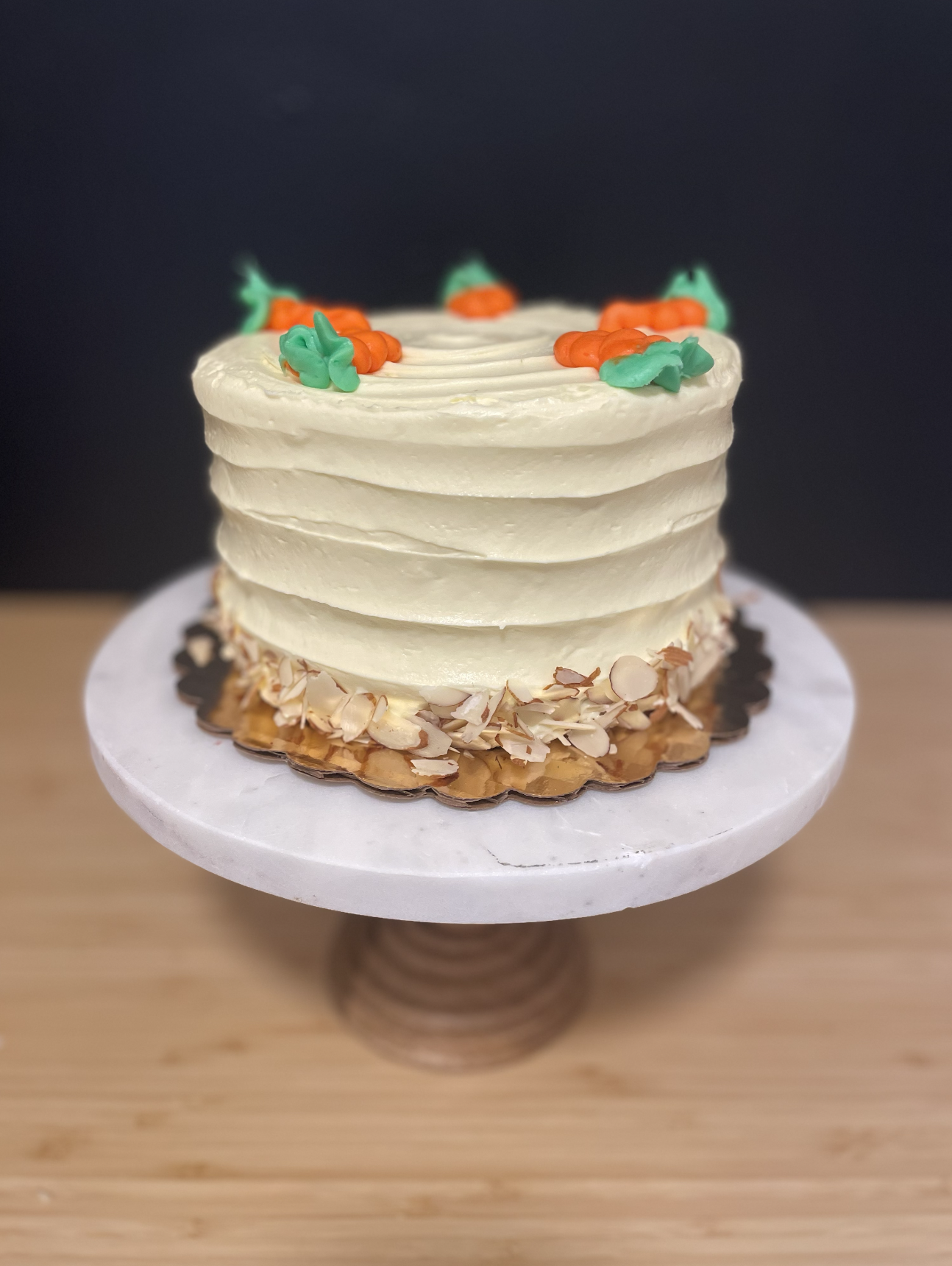 Gluten Free Layered birthday cake with white frosting, topped with colorful carrot-shaped decorations, and almond flakes around the base, on a white cake stand.