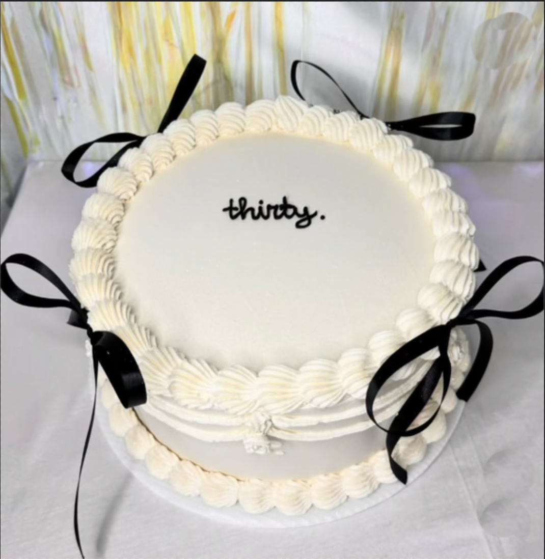 Gluten Free Custom Cakes Round birthday cake with white frosting, decorated with piped white rosettes and black ribbons, with