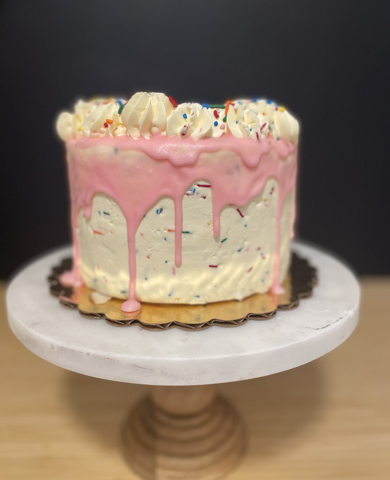 Gluten Free Cakes in Salt Lake City A tall birthday cake with white frosting speckled with colorful sprinkles, topped with white whipped cream swirls and more sprinkles, and pink dripping glaze, on a white cake stand.