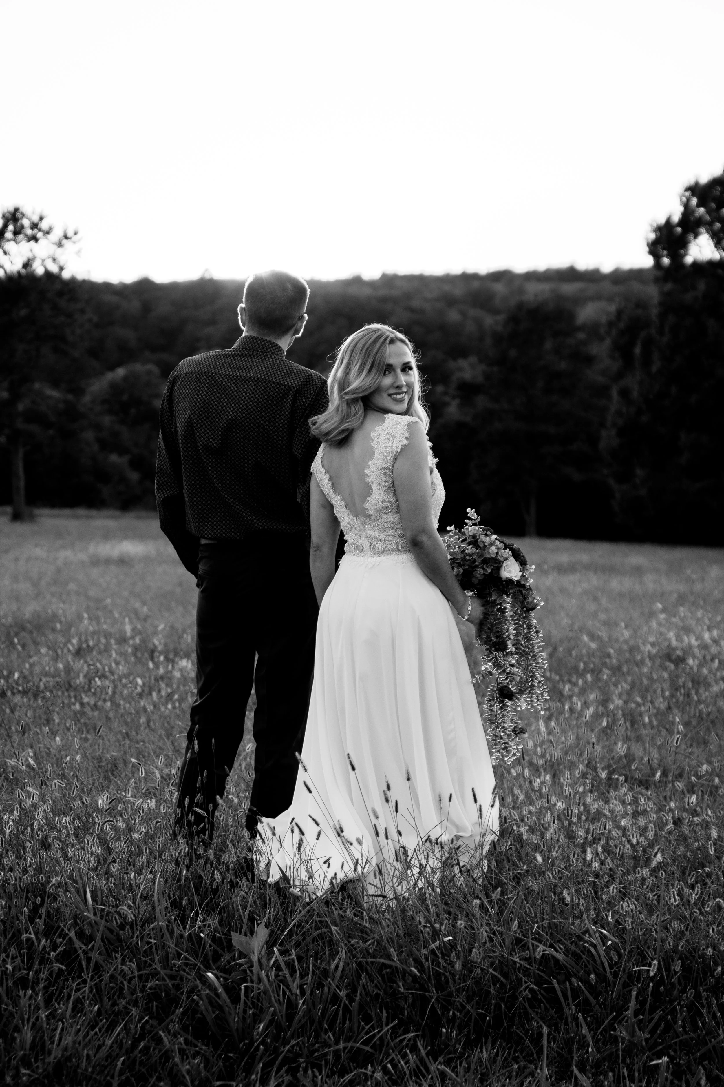 A black-and-white photo of a bride and groom standing in a field, with the bride turning to smile at the camera, holding a bouquet of flowers, while the groom faces away.