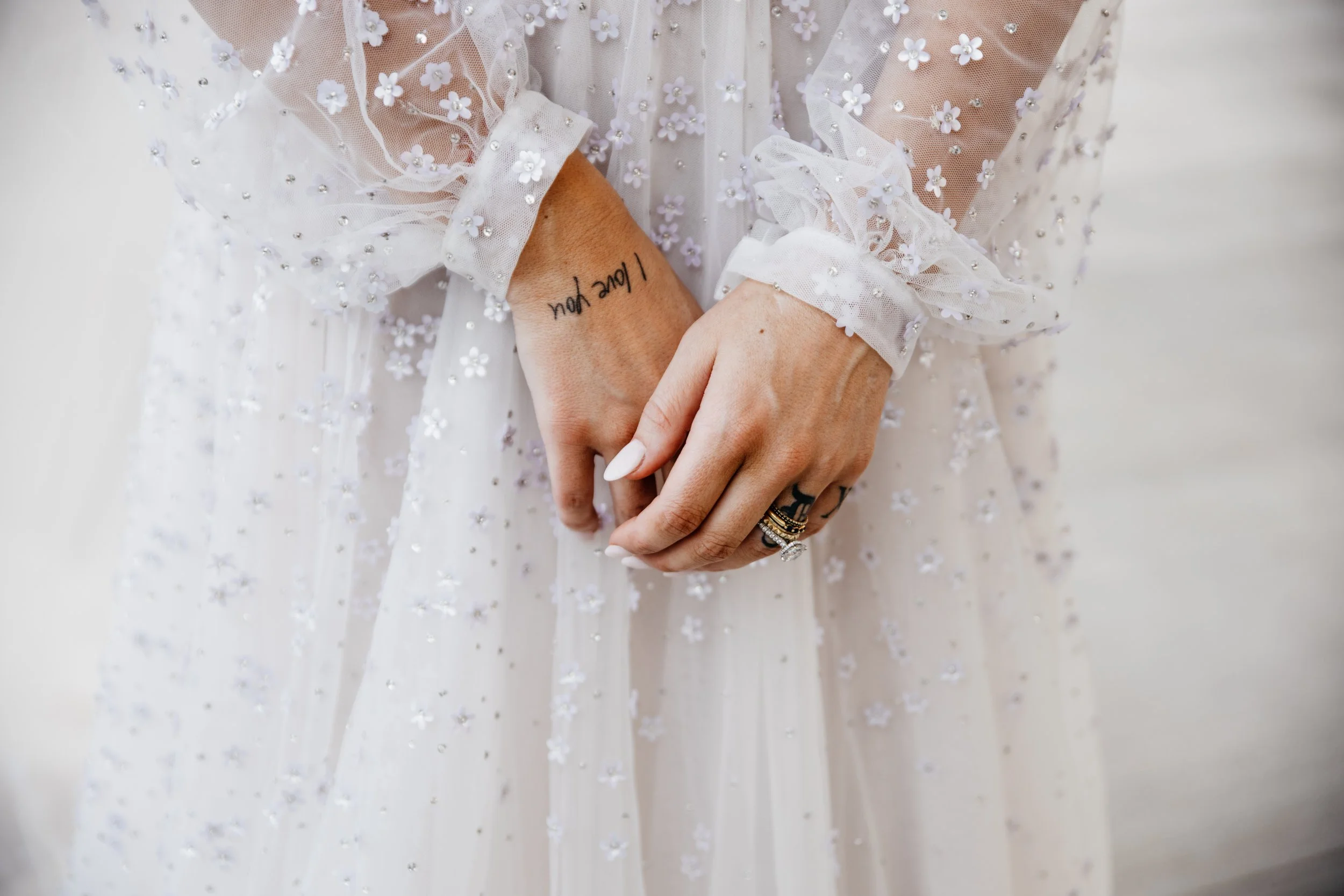 Close-up of a brides’s hands with rings, wearing a sheer, embroidered dress, with the tattoo 'Love you' on her wrist. A thoughtful photograph from Keri-Ann Gordon, International photographer