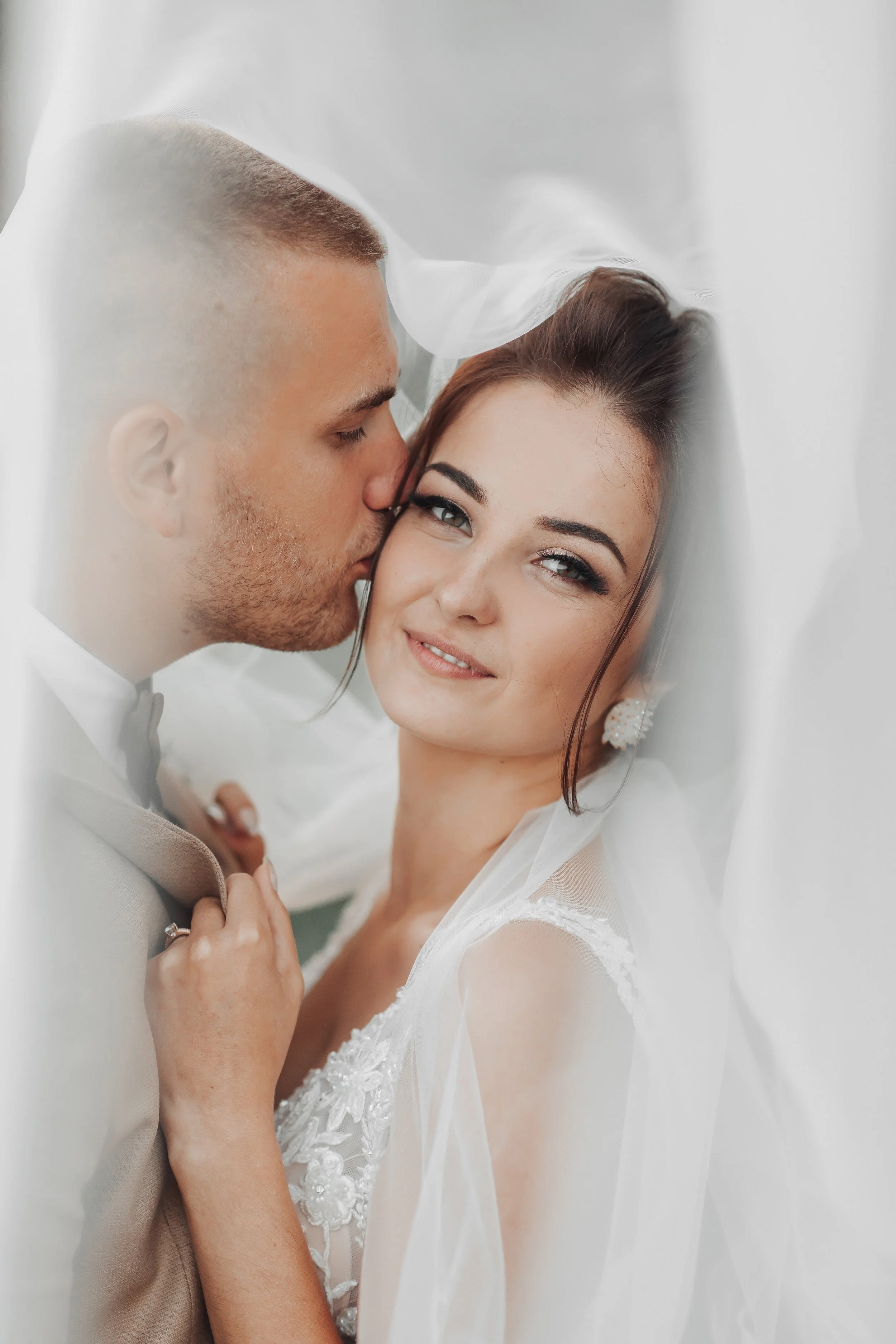 Bride and groom on wedding day, with the groom kissing the bride's temple, bride smiling softly, in soft focus with white veil and dress, photographed by KA Gordon, Kansas's premier wedding photographer