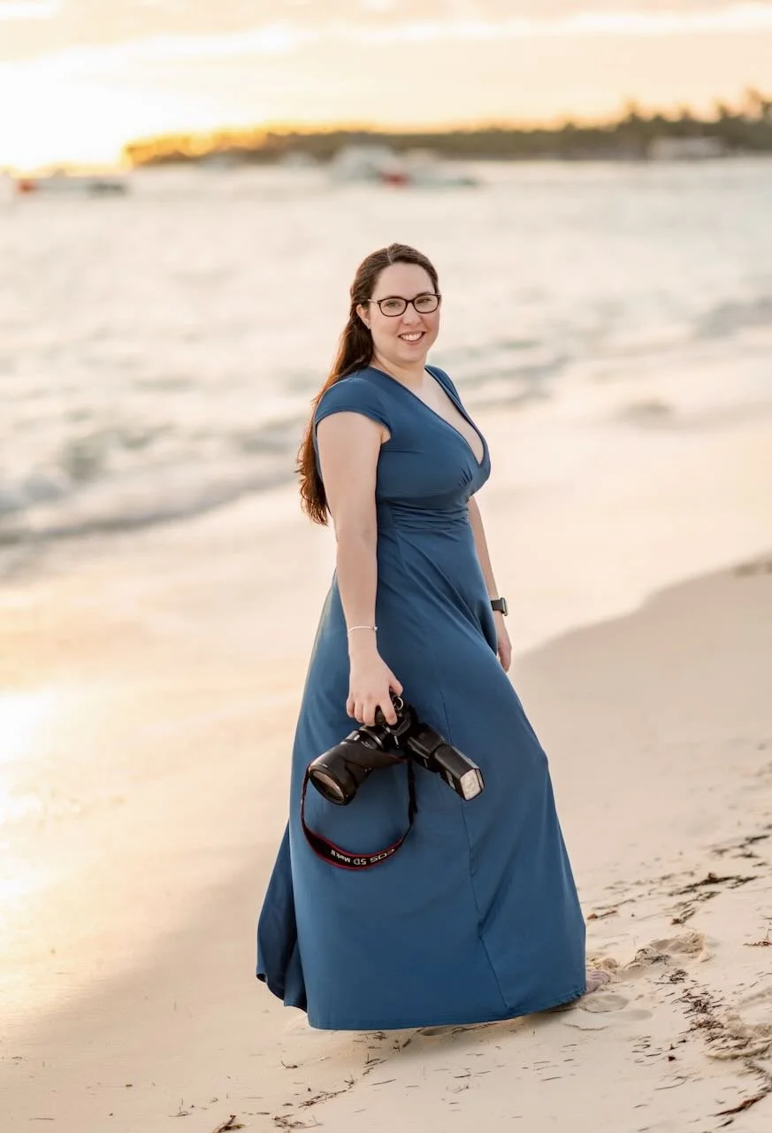 Keri-Ann Gordon, destination wedding photographer, wearing glasses and a blue dress standing barefoot on the beach at sunset, holding a camera with flash.