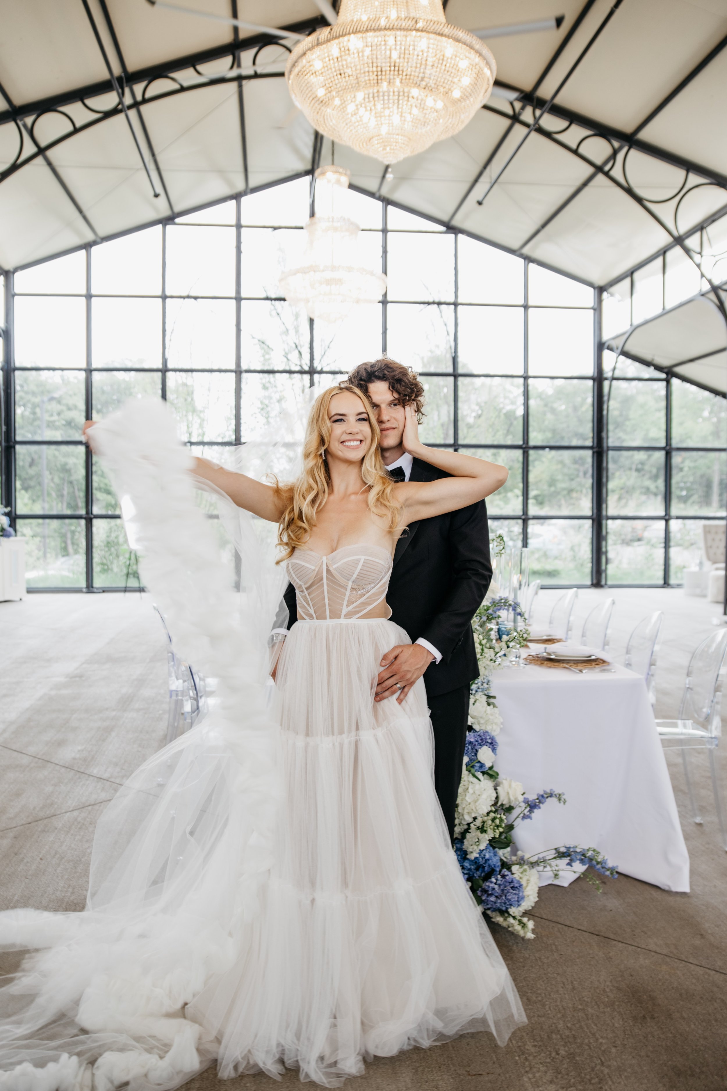 A bride and groom in wedding attire at their destination wedding posing inside a glass-paneled venue with chandeliers, with a decorated table and floral arrangements in the background