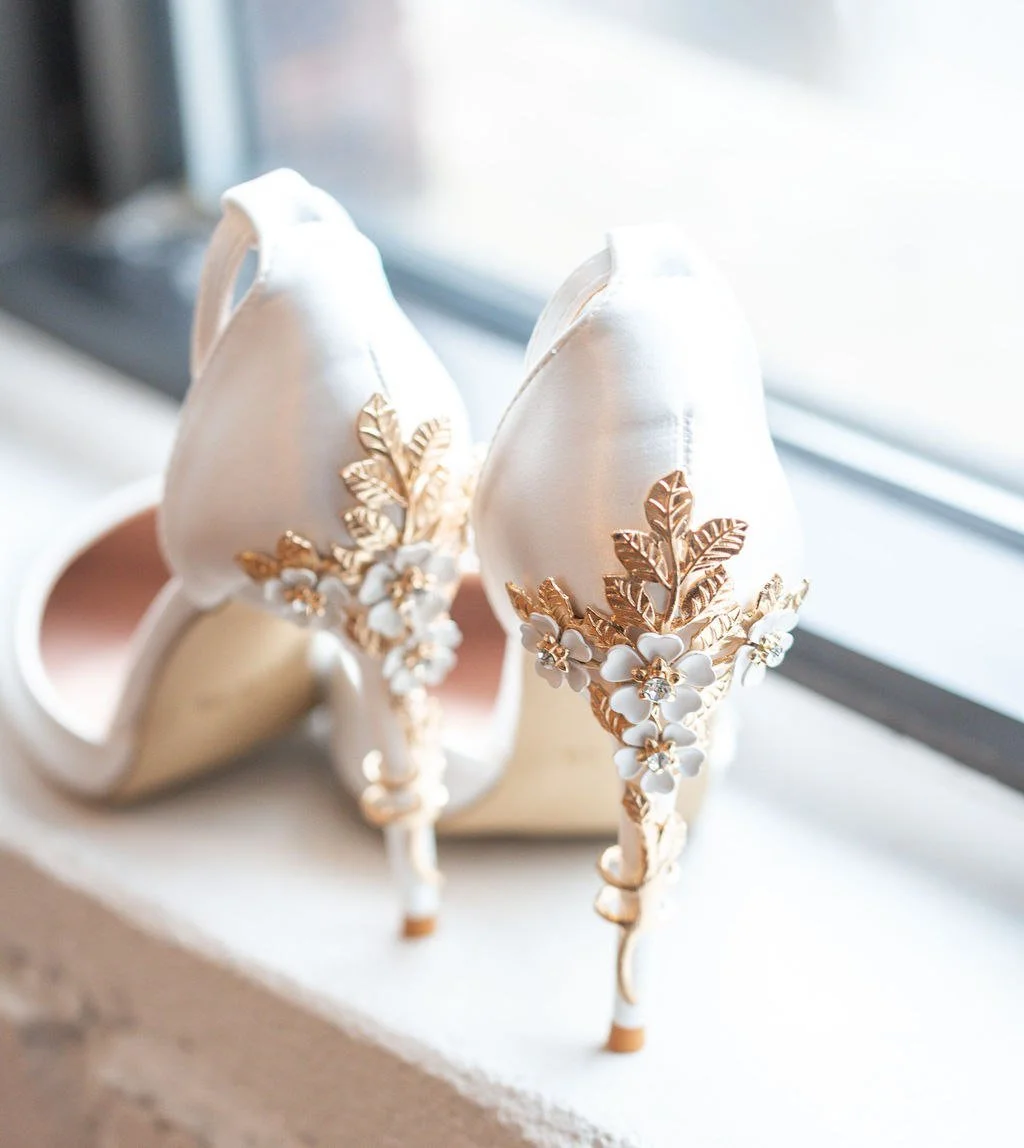 Keri-Ann Gordon photographs a pair of Elegant white high-heeled shoes with gold floral embellishments and white flowers, placed near a window.