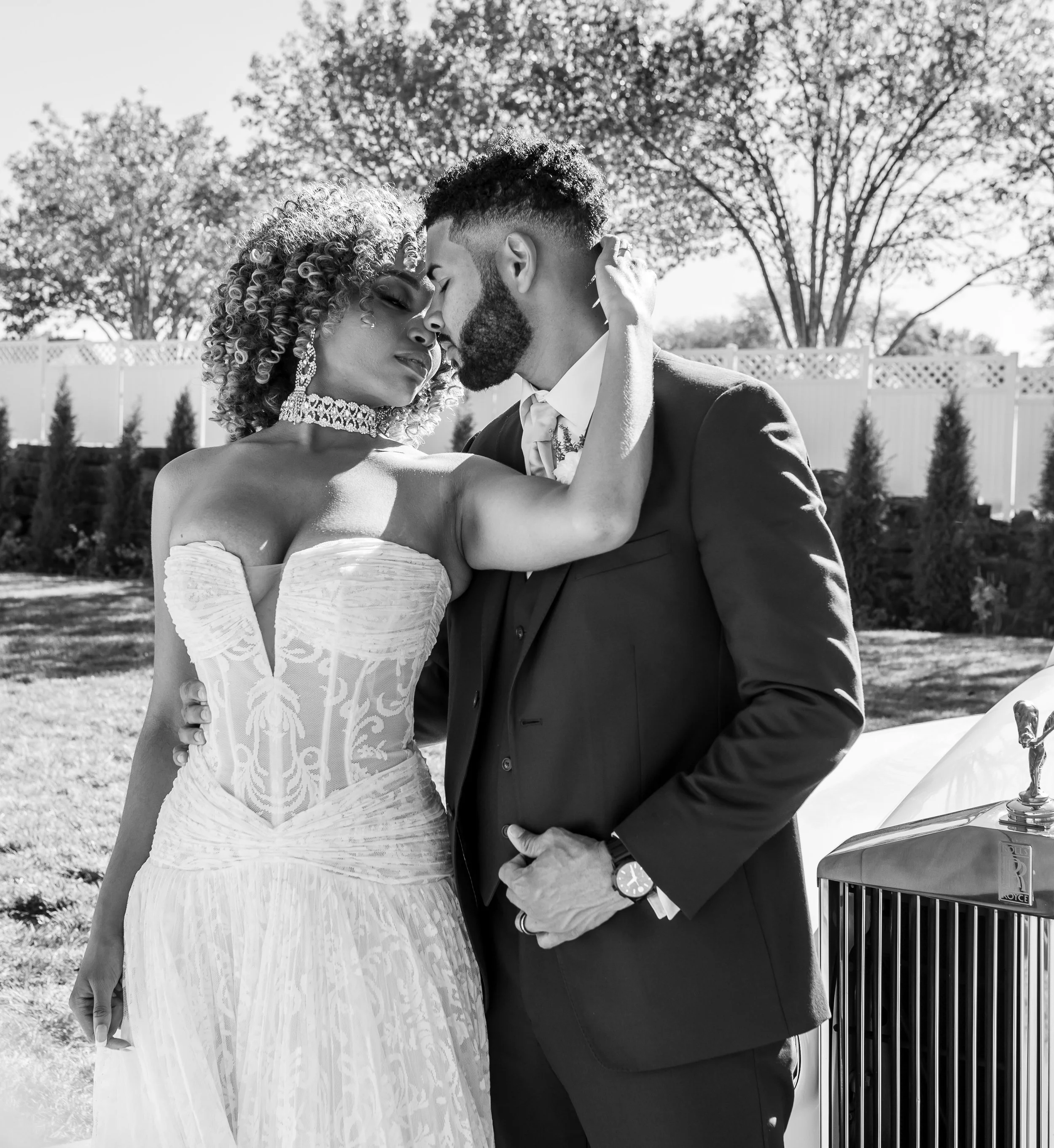 Black and white photograph of a bride and groom embracing outdoors during daytime, with trees and a white fence in the background. The bride wears an off-shoulder wedding dress, and the groom wears a suit. They have their foreheads and noses touching, with eyes closed.