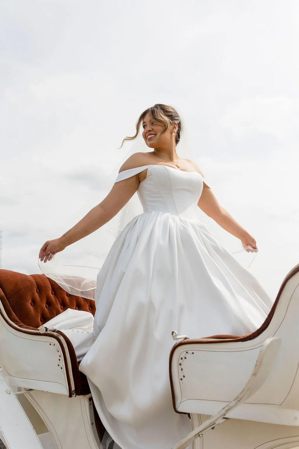 A bride in a white wedding dress standing outdoors, smiling, with her arm outstretched, against a cloudy sky. A stunning image captured by international wedding photographer KA Gordon