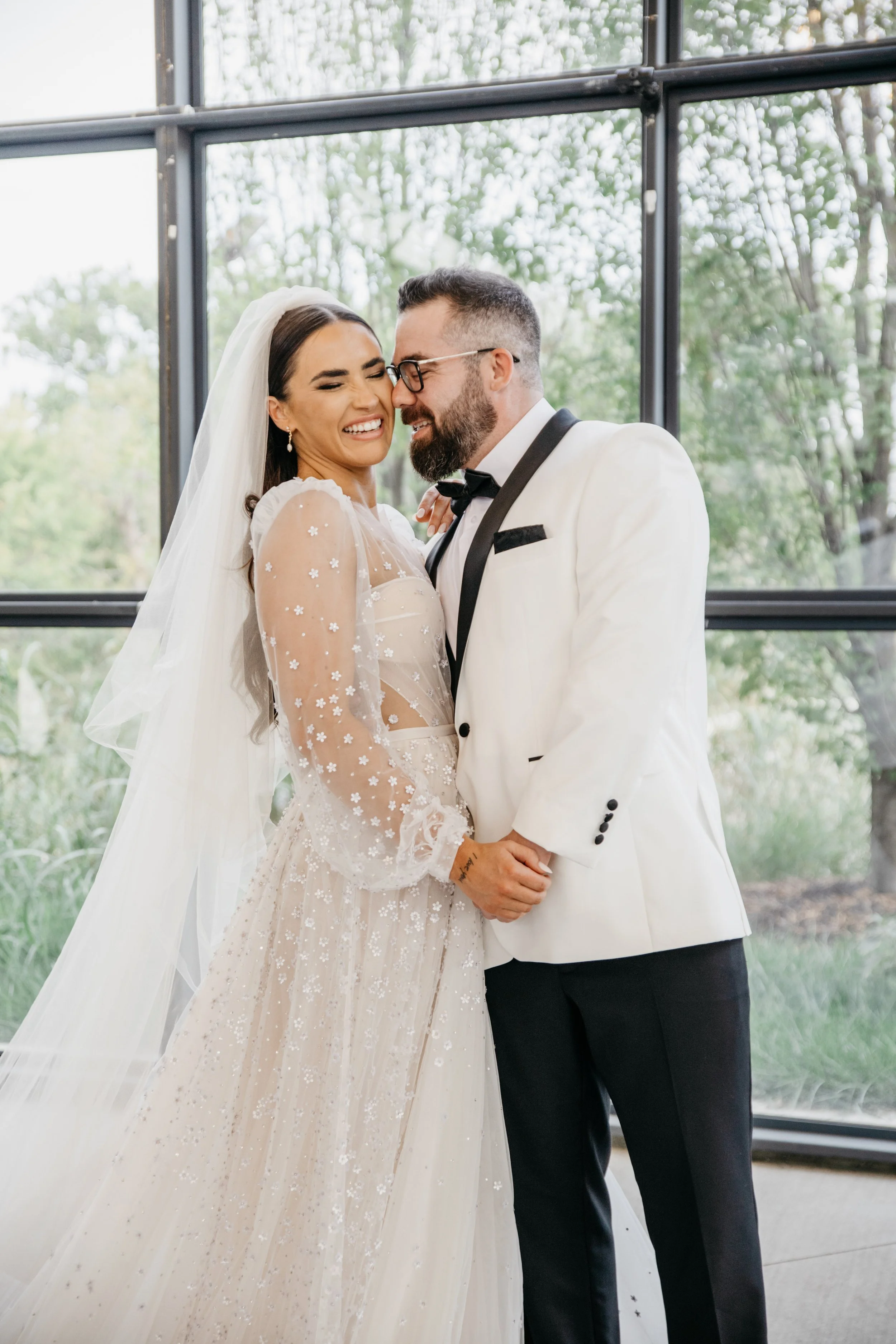 A bride in a white wedding dress with floral embroidery and a veil, and a groom in a white tuxedo with black lapels, share a joyful moment indoors with large glass windows and greenery outside at her international destination wedding