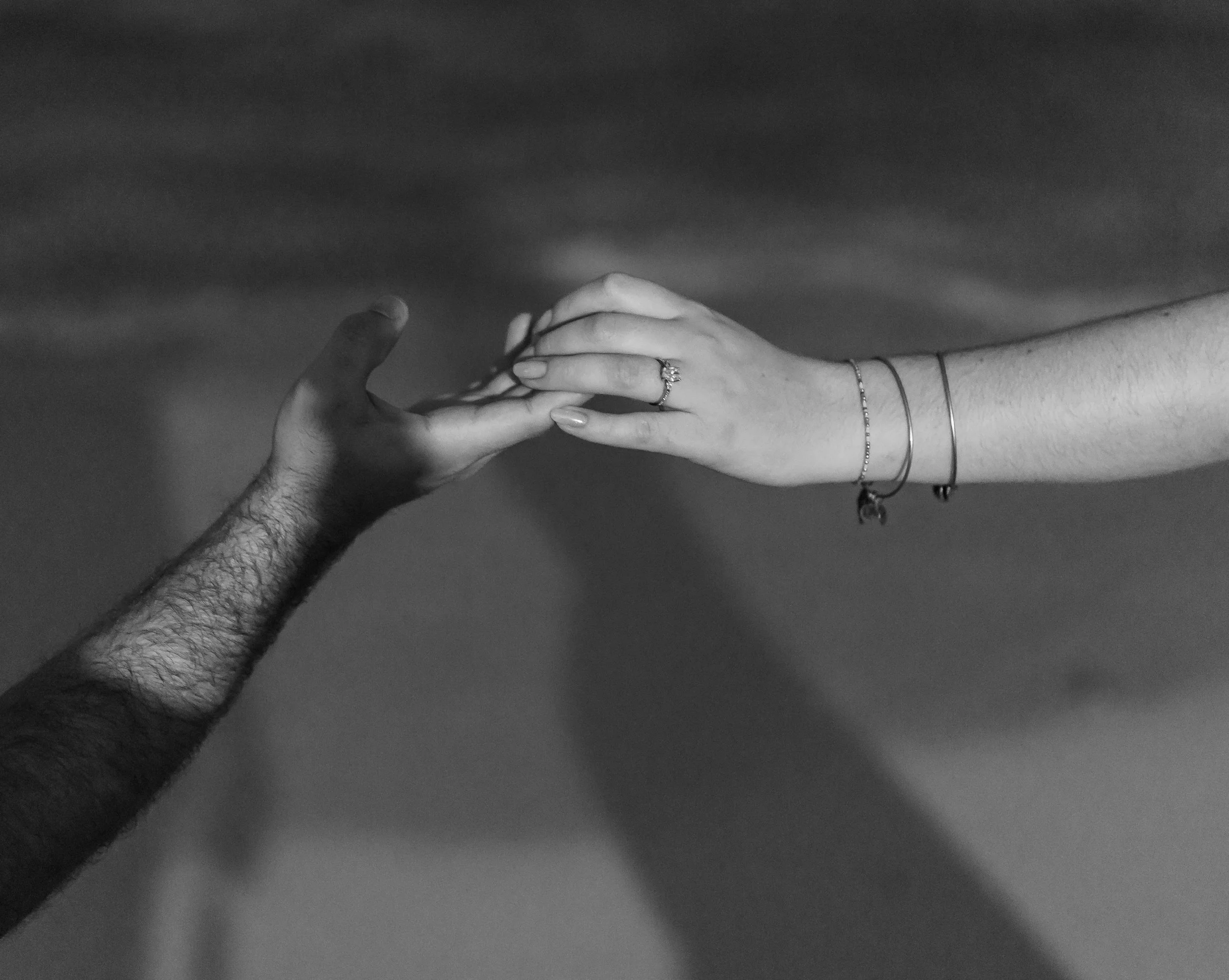 Two hands reaching out and touching each other, one with a wedding ring and bracelets, and the other with arm hair, against a plain background.