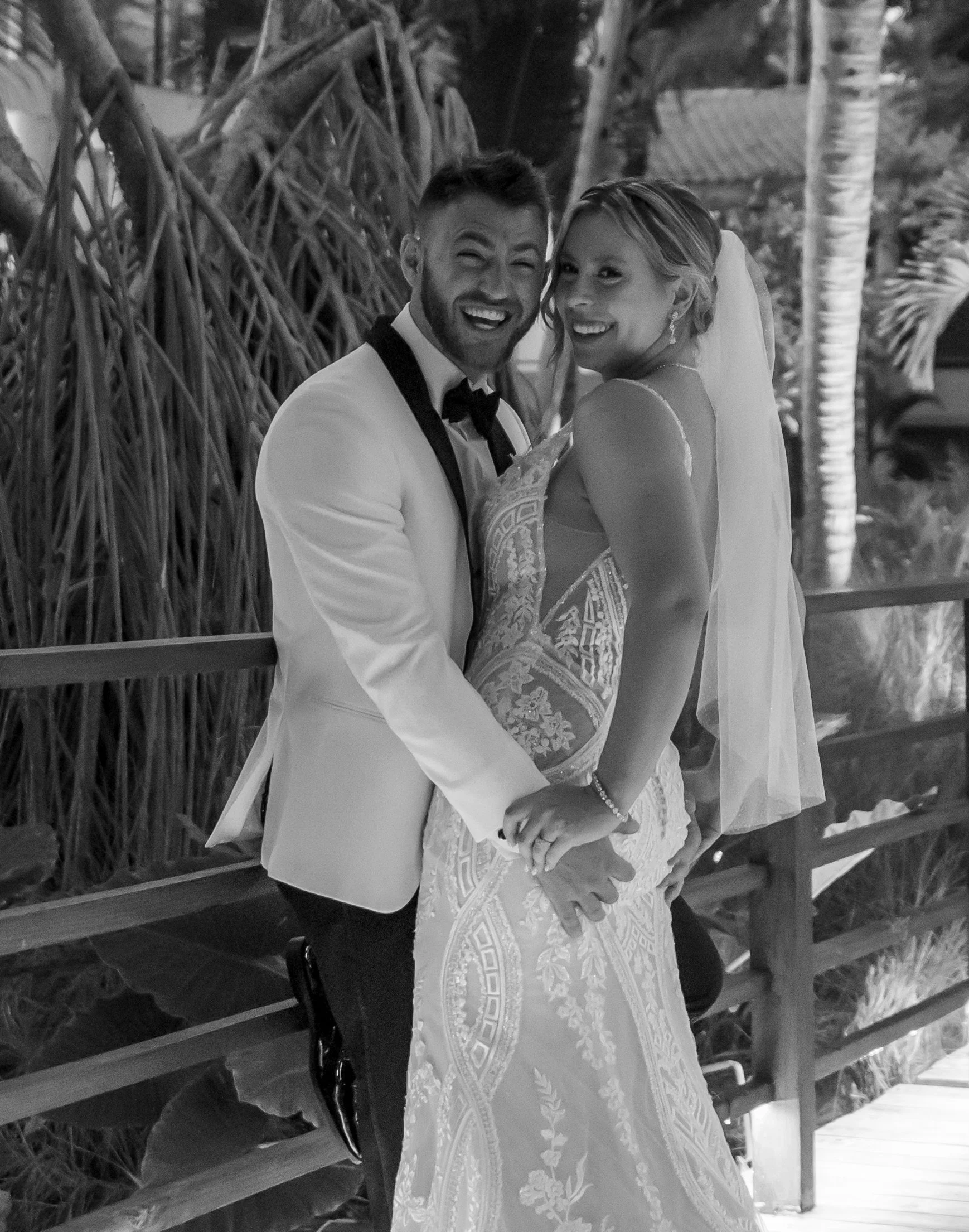 Happy bride and groom in wedding attire embrace on a wooden bridge with lush tropical plants in the background.