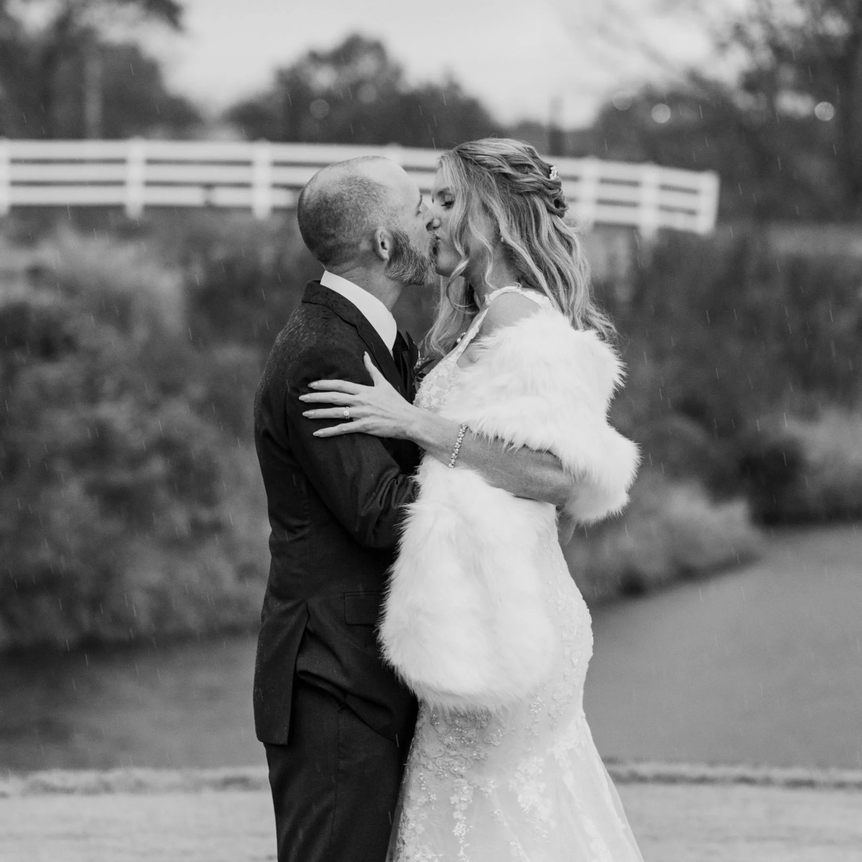 A black and white photo of a bride and groom kissing outdoors in the rain, with a white fence and trees in the background.