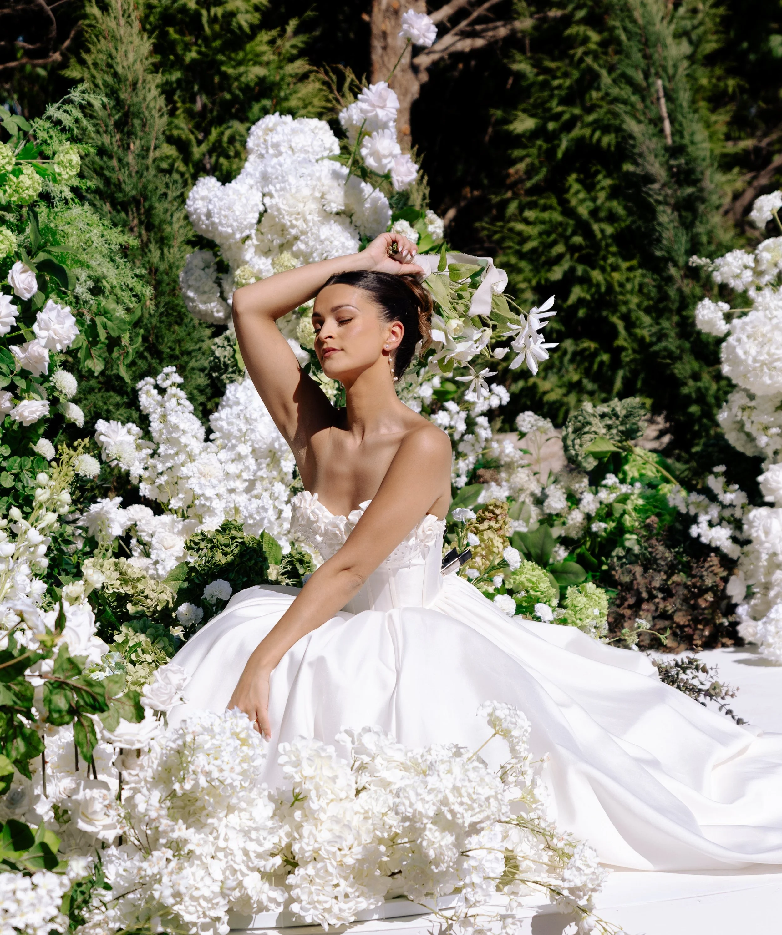 A woman in a white wedding dress sitting among white flowers and greenery with her eyes closed and one arm raised above her head. A dreamy photograph Captured by international wedding photography KA Gordon