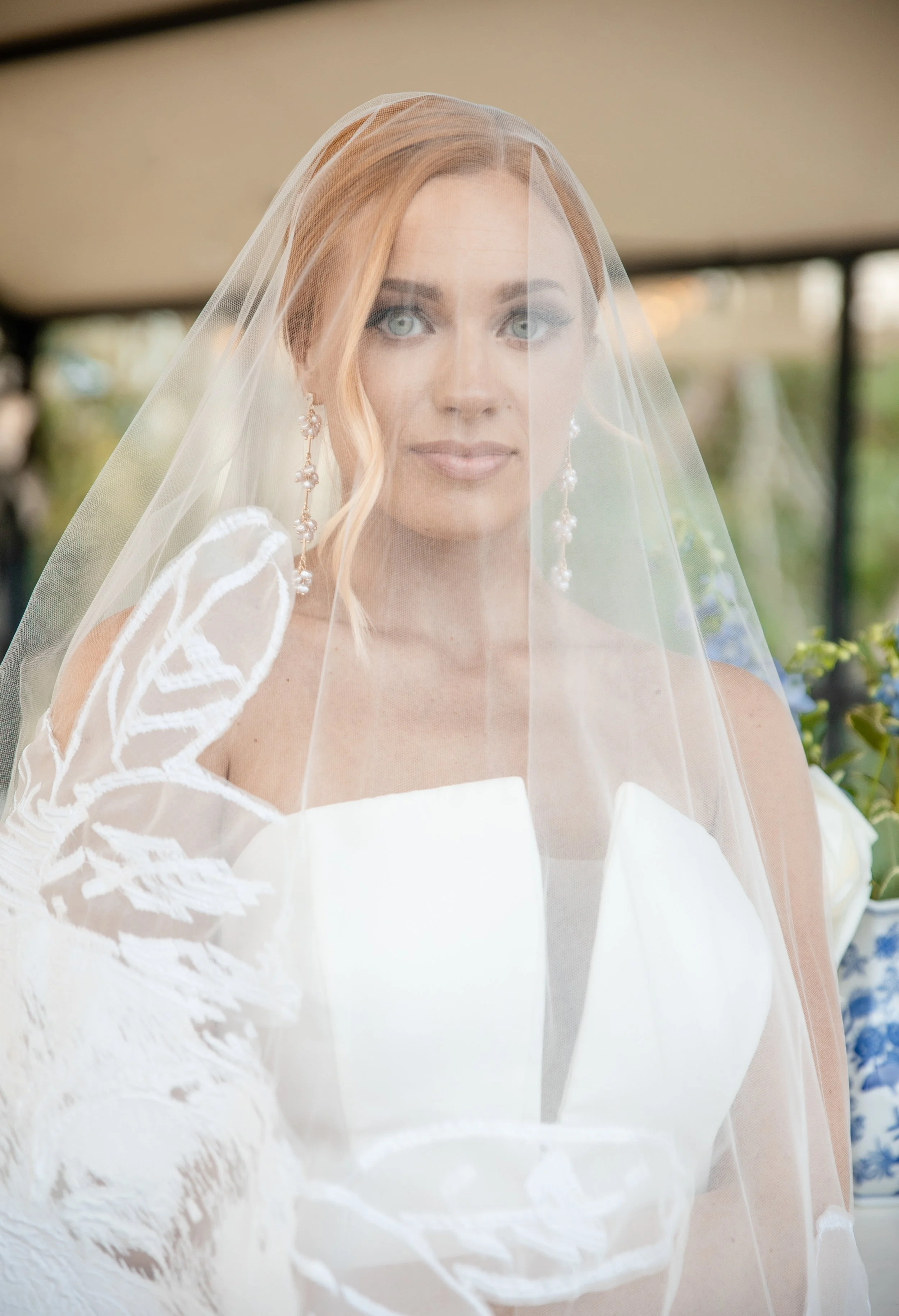 A bride wearing a white wedding dress, veil, and long earrings, holding gloves with lace details, indoors with a blurred background. Kansas wedding photographer