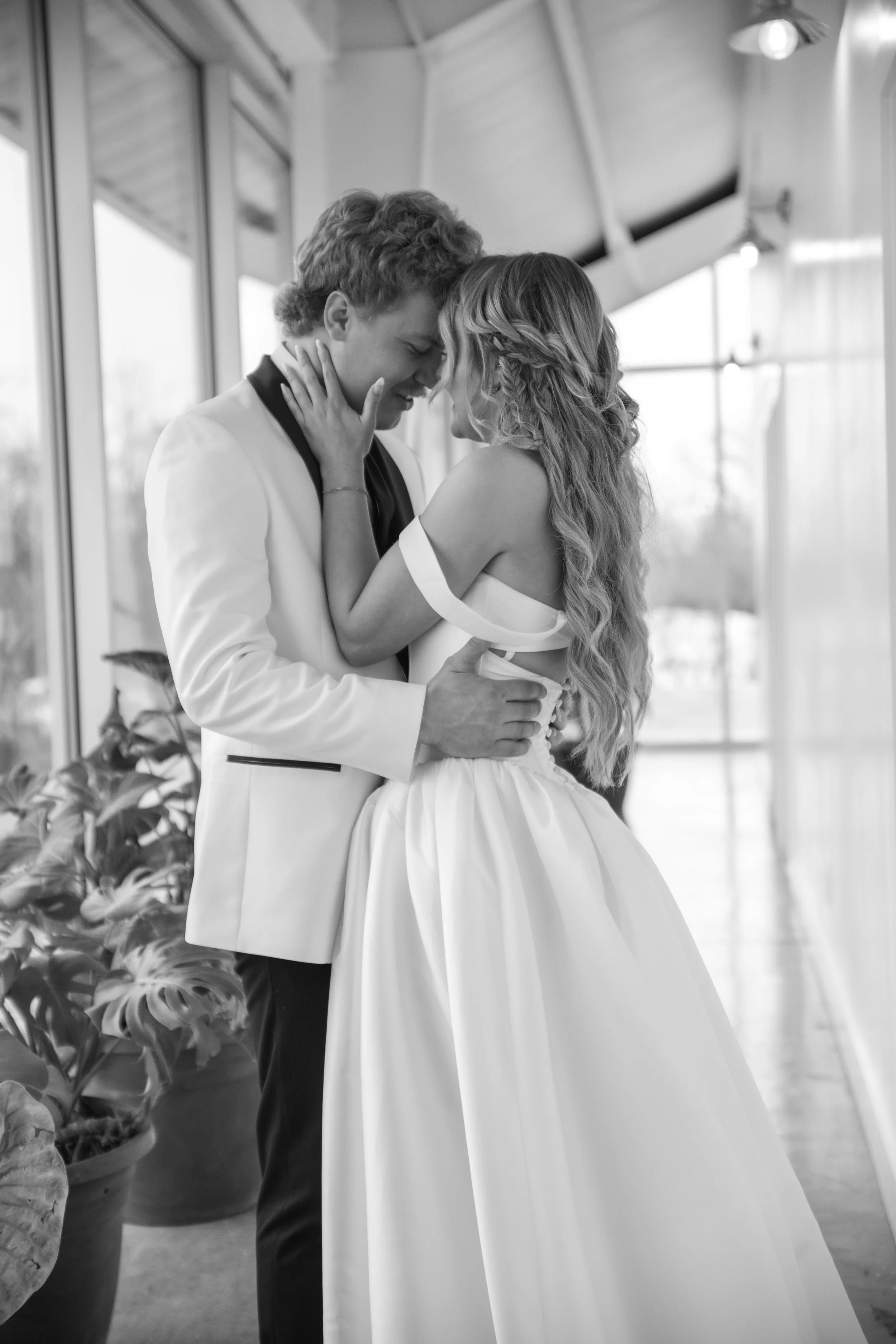 A black and white photo of a couple, with their foreheads touching and eyes closed, embracing tenderly indoors and dancing to the first dance at their international wedding
