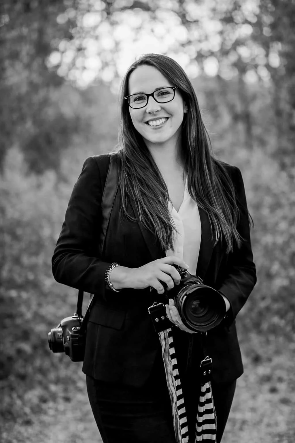 A smiling Keri-Ann Wedding Photographer, with long hair and glasses holding her professional camera at an outdoor wedding