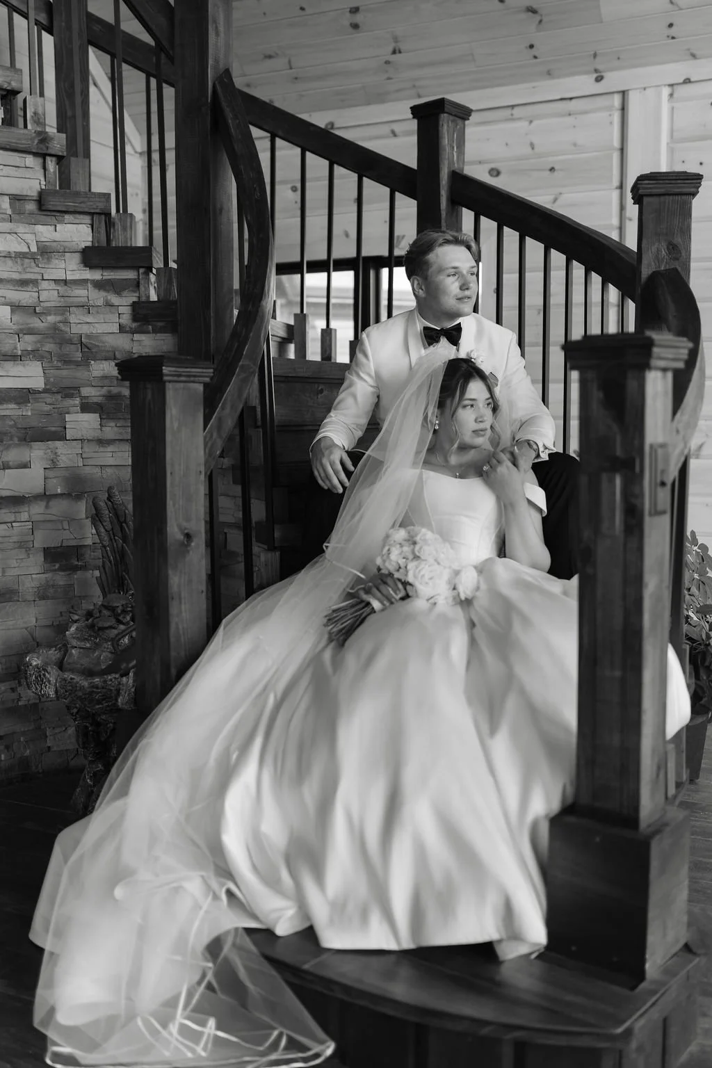 A bride and groom sitting on a staircase indoors, with the bride holding a bouquet of flowers, and both dressed in wedding attire.