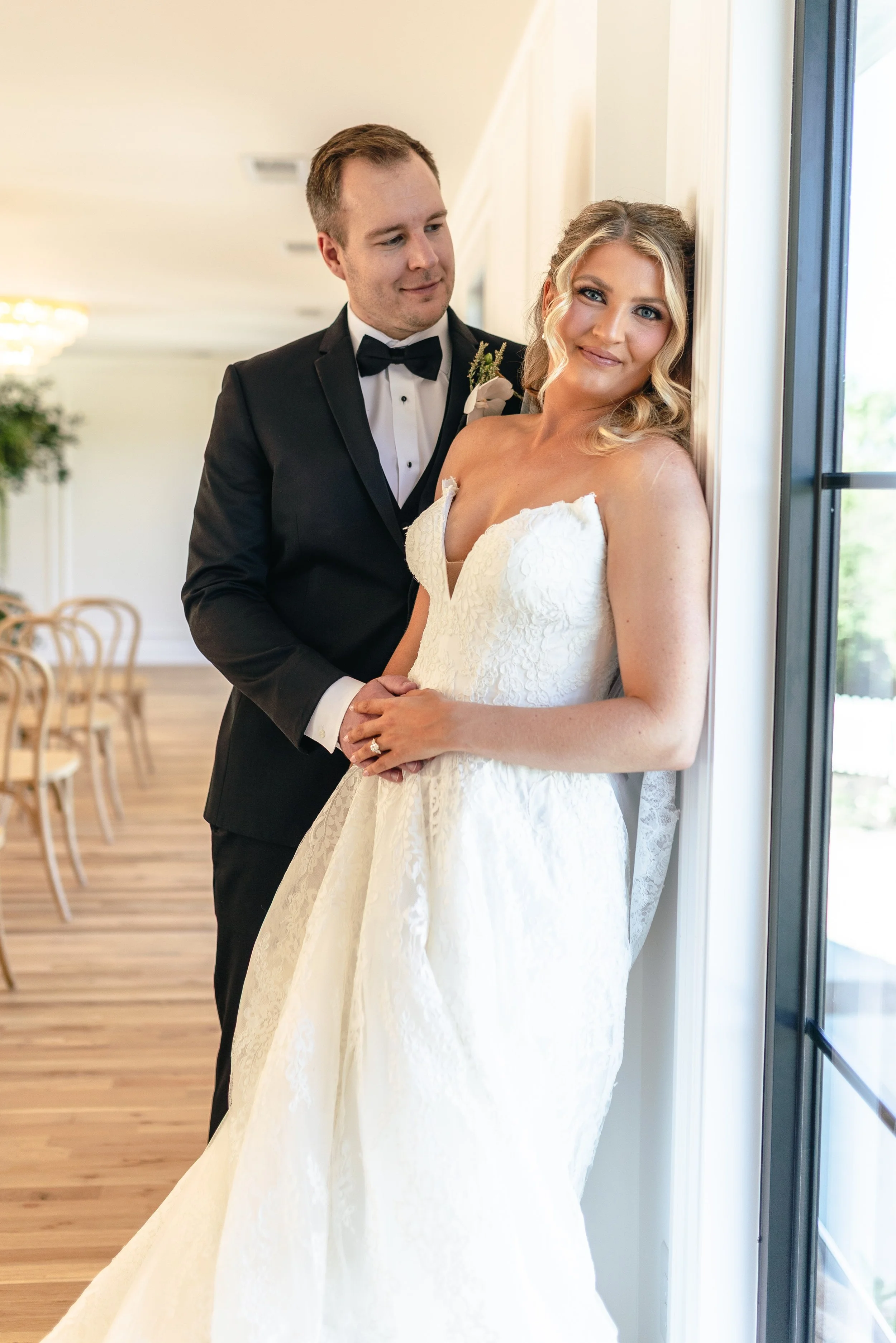 A bride and groom on their wedding day, standing near a window with sunlight. The bride is wearing a strapless white wedding dress, and the groom is in a black tuxedo with a bow tie. The groom is holding the bride's hands, and they are looking at each other.