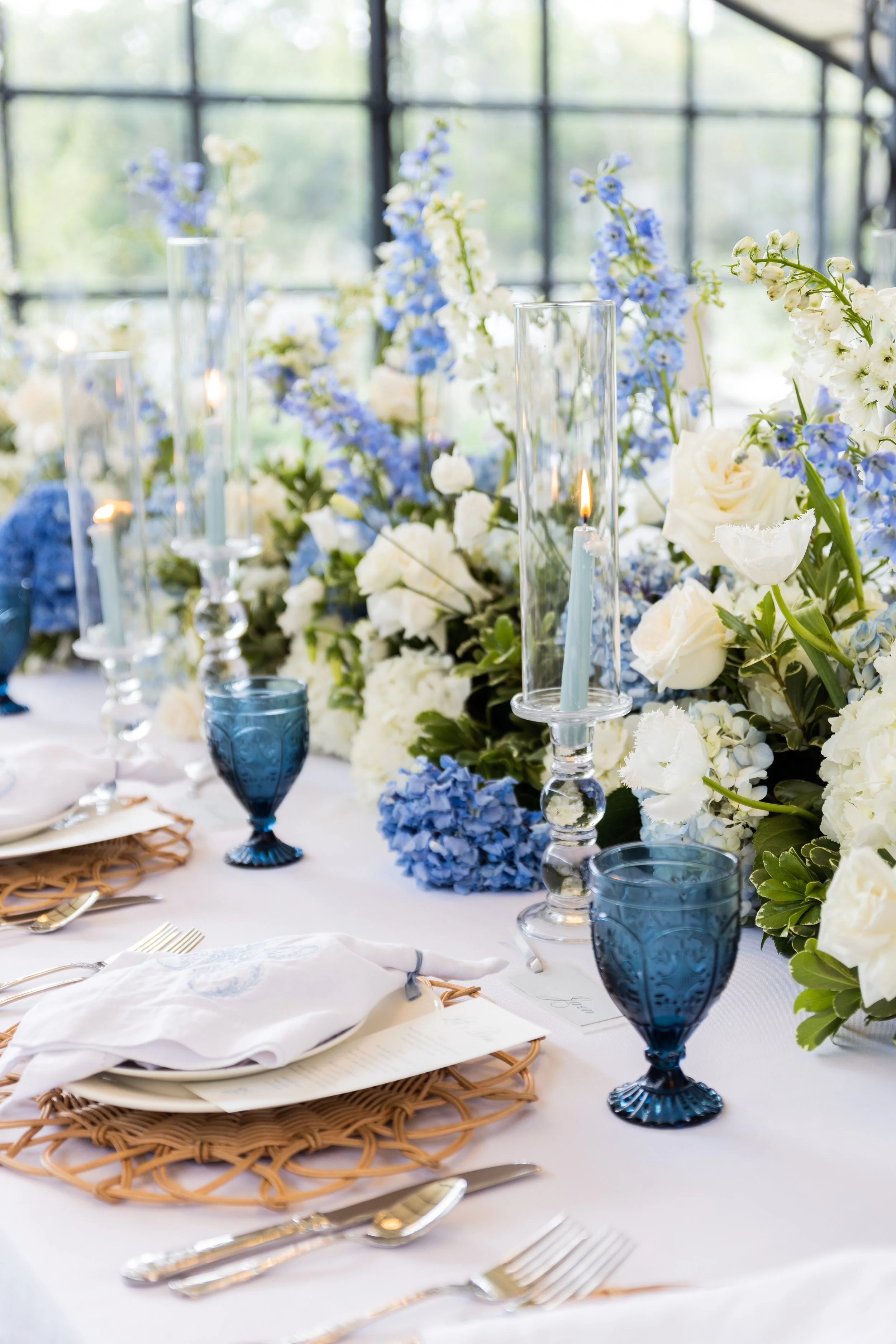 Elegant table setting with white napkins, gold flatware, blue glassware, tall glass candle holders, and a lush centerpiece of white and blue flowers