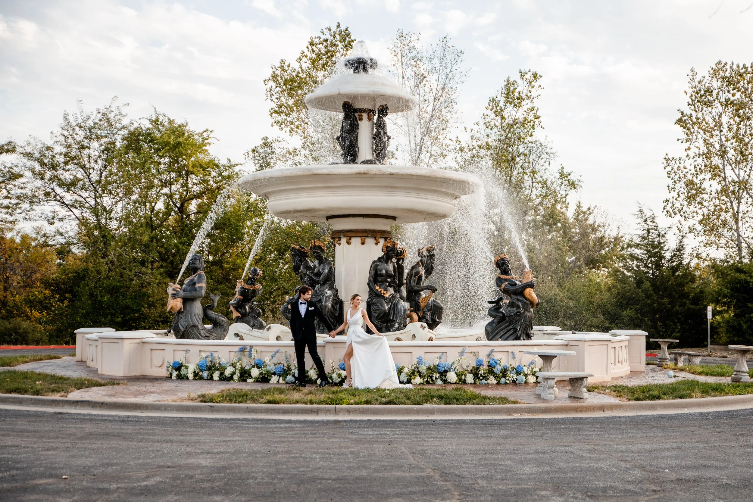A newlywed couple in formal wedding attire standing in front of a decorative fountain with water spraying from several statues, surrounded by trees and flowers.