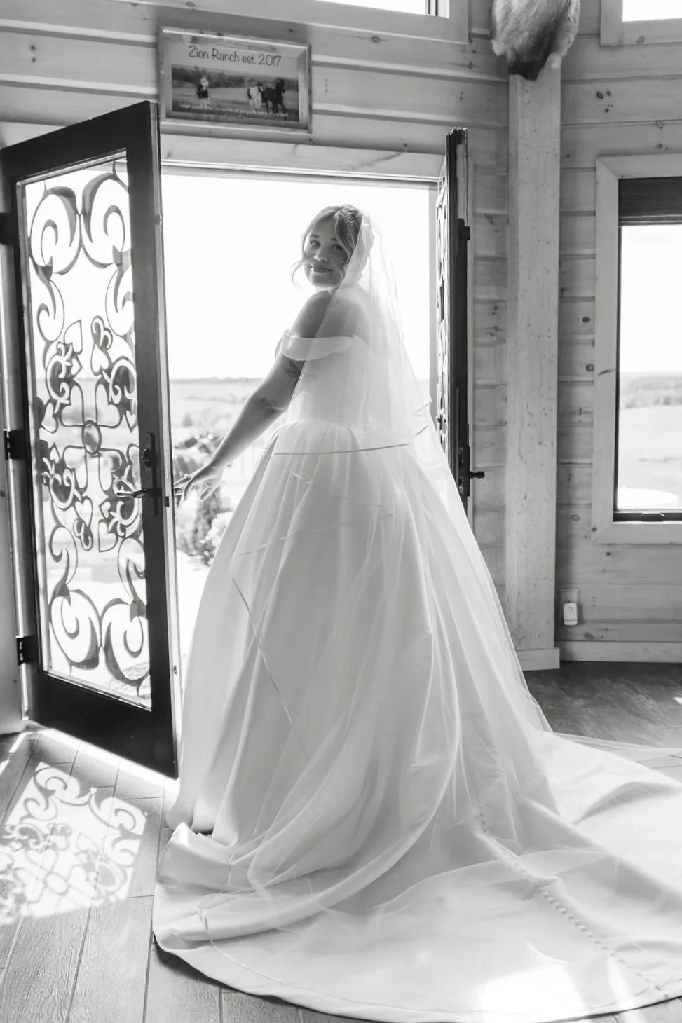 A bride in a wedding dress standing in a doorway, smiling and looking outside. The interior has wooden walls and a window, and a picture frame on the wall reads "Zion Ranch est. 2017."  Photographed by Keri-Ann Gordon editorial wedding photographer