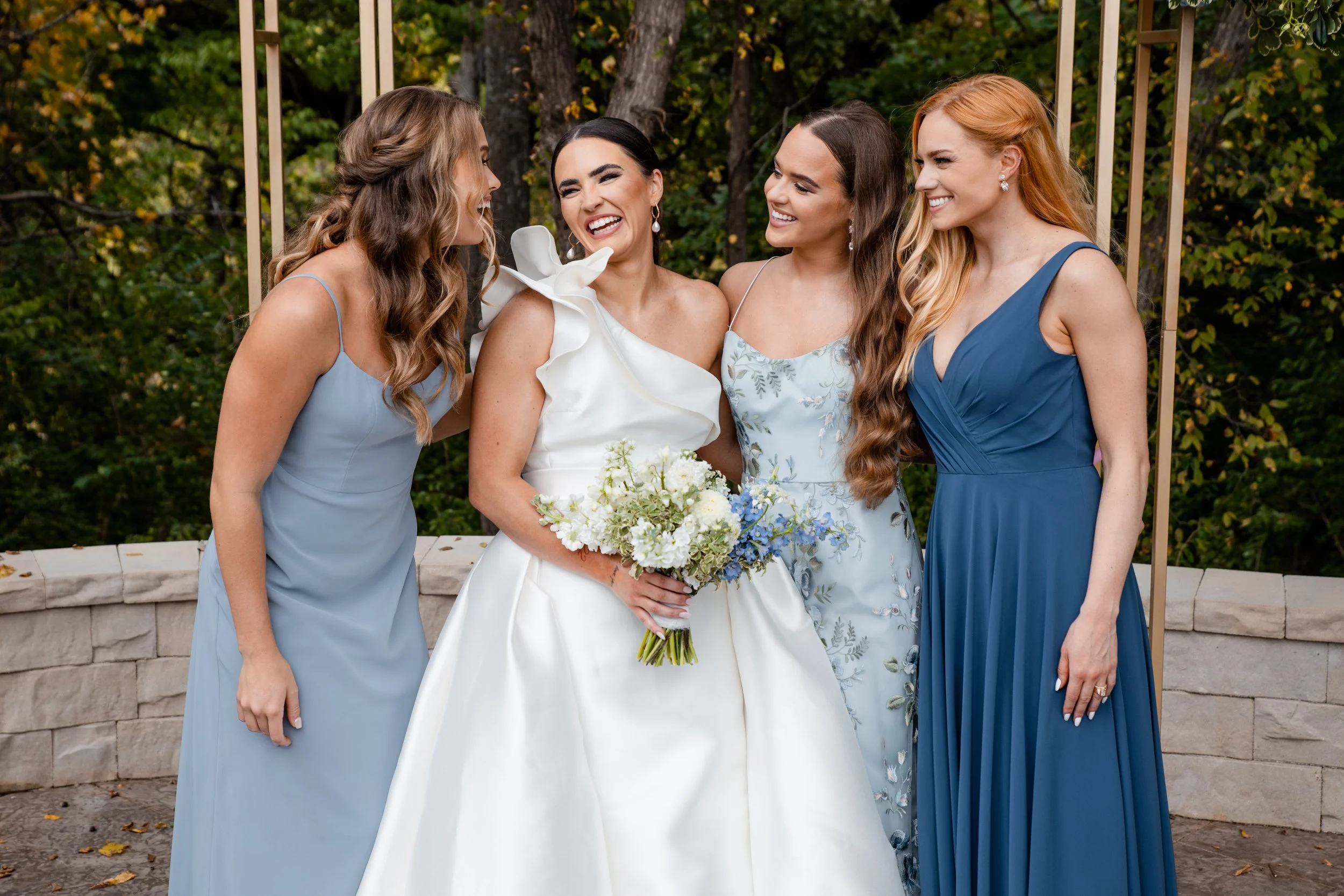 a bride in a white wedding gown holding a bouquet, standing outdoors surrounded by her three bridesmaids dressed in blue, smiling and laughing together at the bride's Kansas City wedding