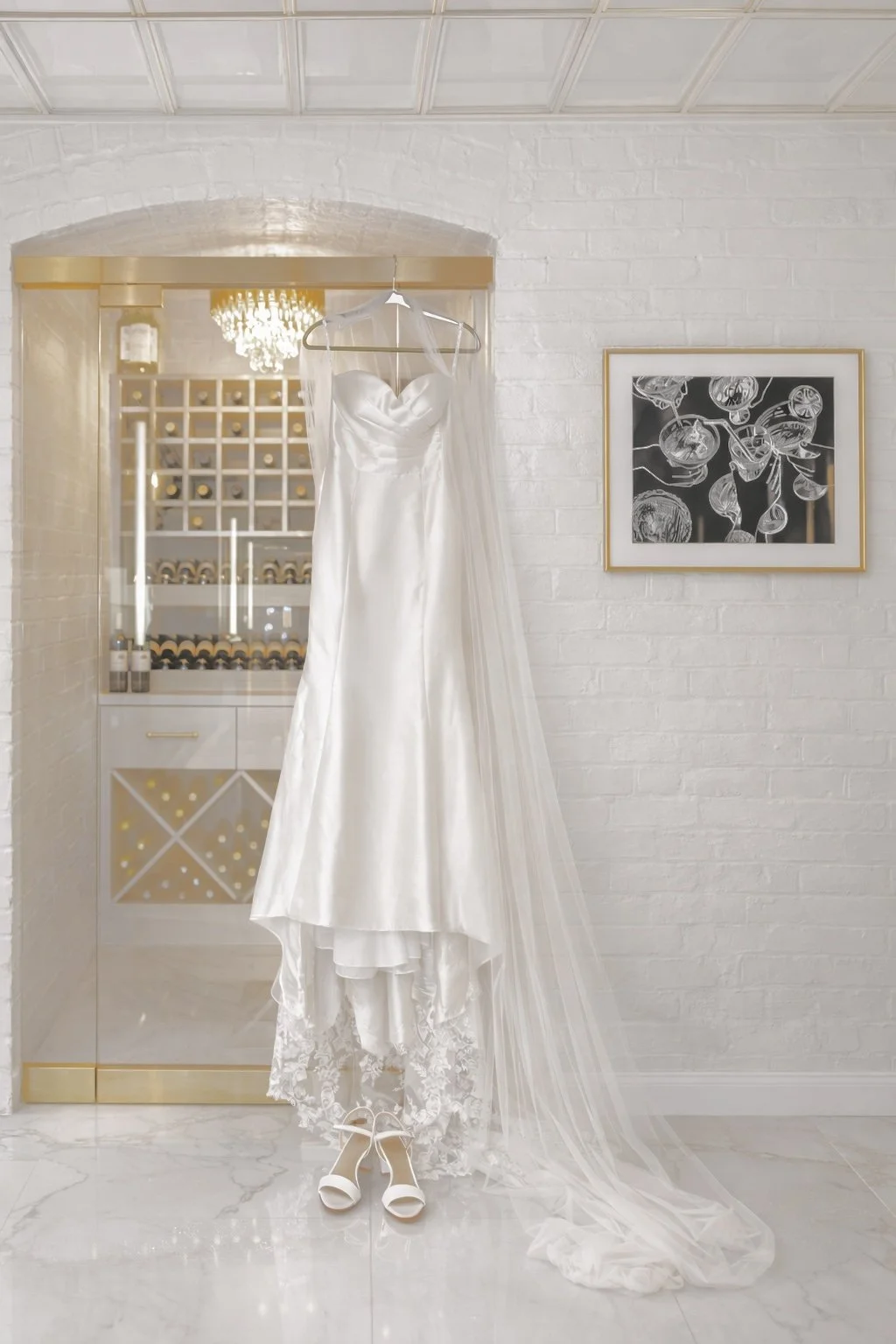White wedding dress with veil hanging in front of a wine display cabinet, with white shoes at the bottom, and a framed black-and-white floral artwork on a white brick wall, destination wedding photographer worldwide