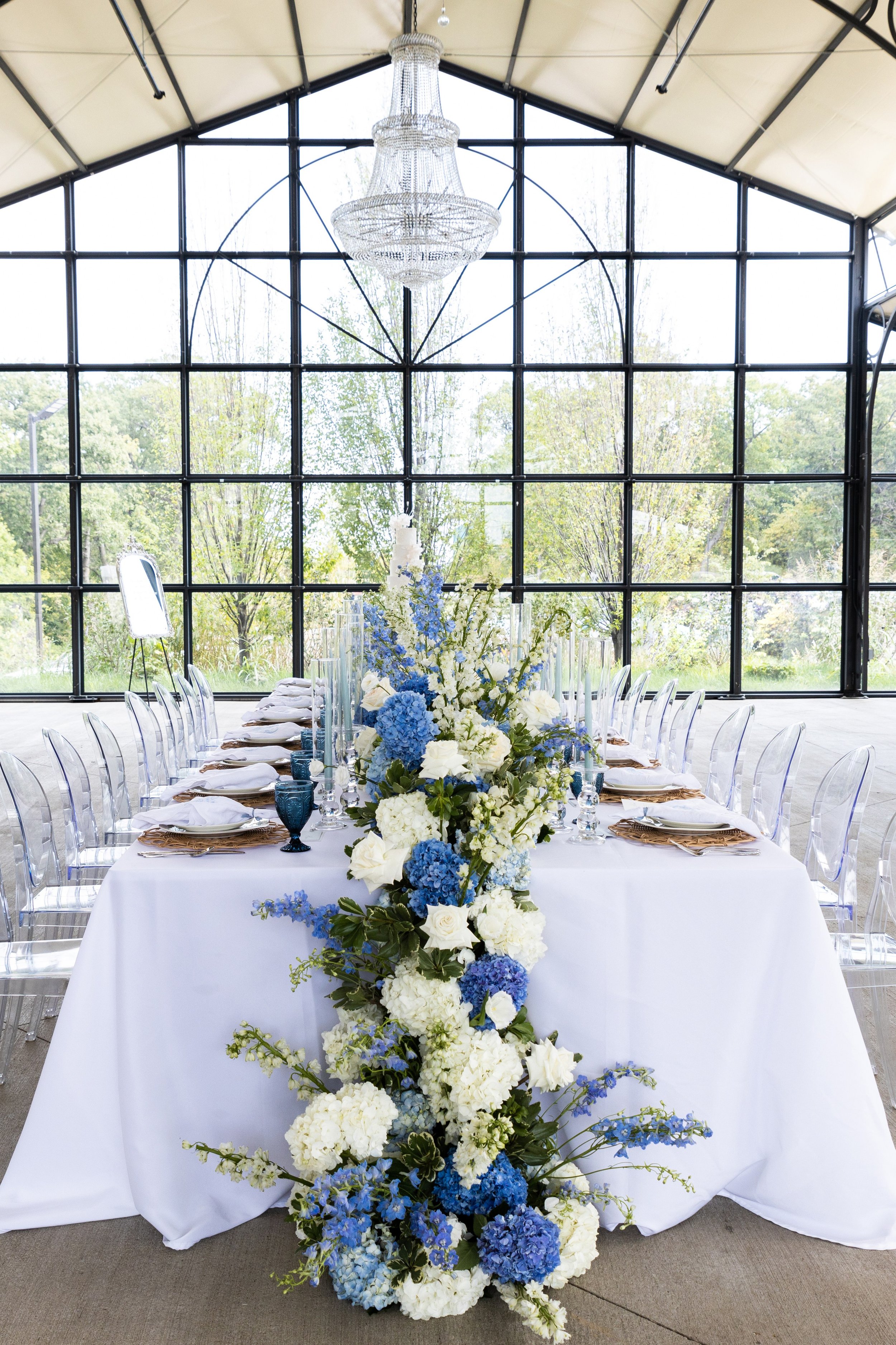Elegant indoor dining setup with a long table, white tablecloth, and clear acrylic chairs. The table is decorated with a cascading floral centerpiece of white and blue flowers, and tall candles. The background features a large window with black framing, allowing natural light to illuminate the space, and a chandelier hanging from the ceiling.