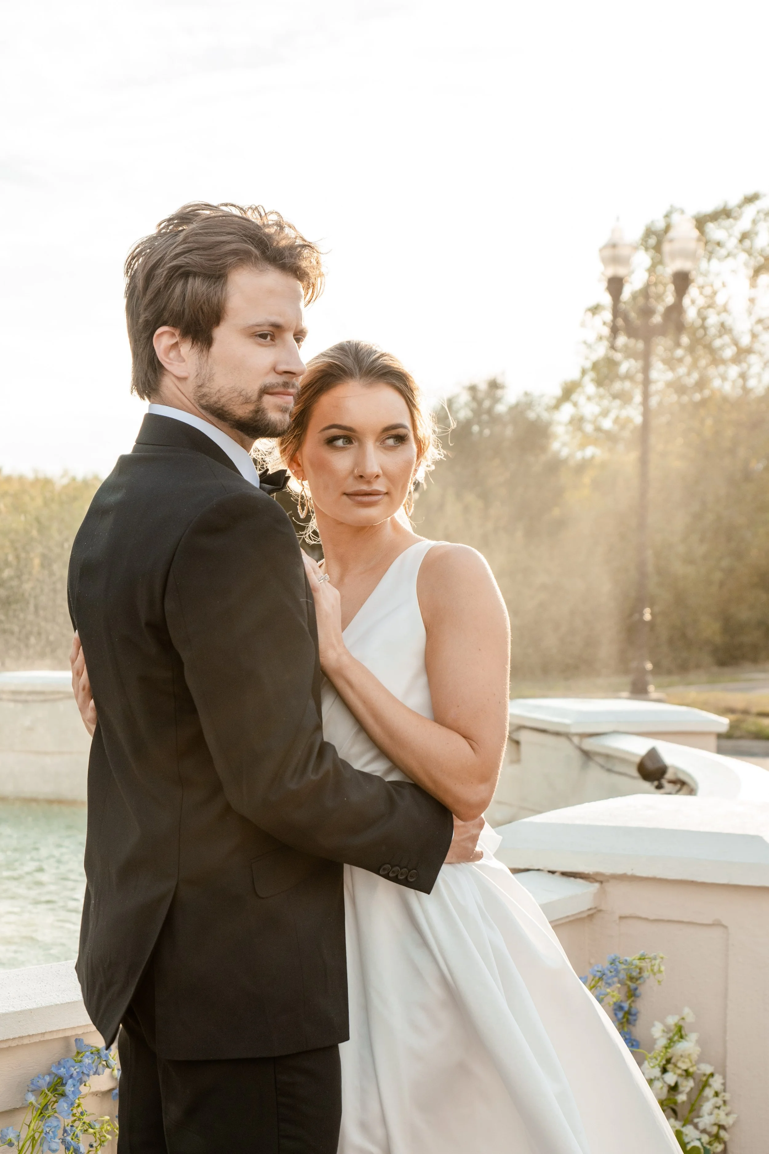 A bride and groom pose for a wedding photo outdoors during sunset, with the woman in a white wedding dress and the man in a black tuxedo.