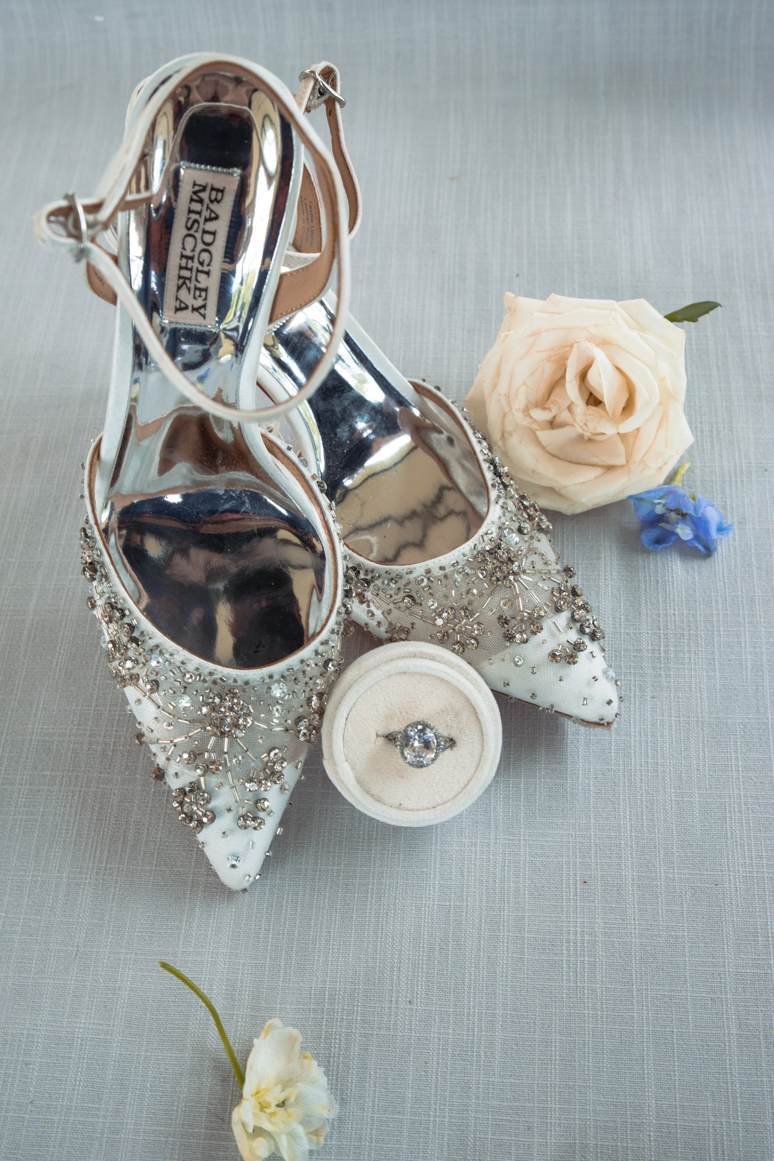 A pair of white, embellished high-heeled shoes with rhinestones, a white ring in a velvet box, a white rose, and a small blue flower on a light gray surface.