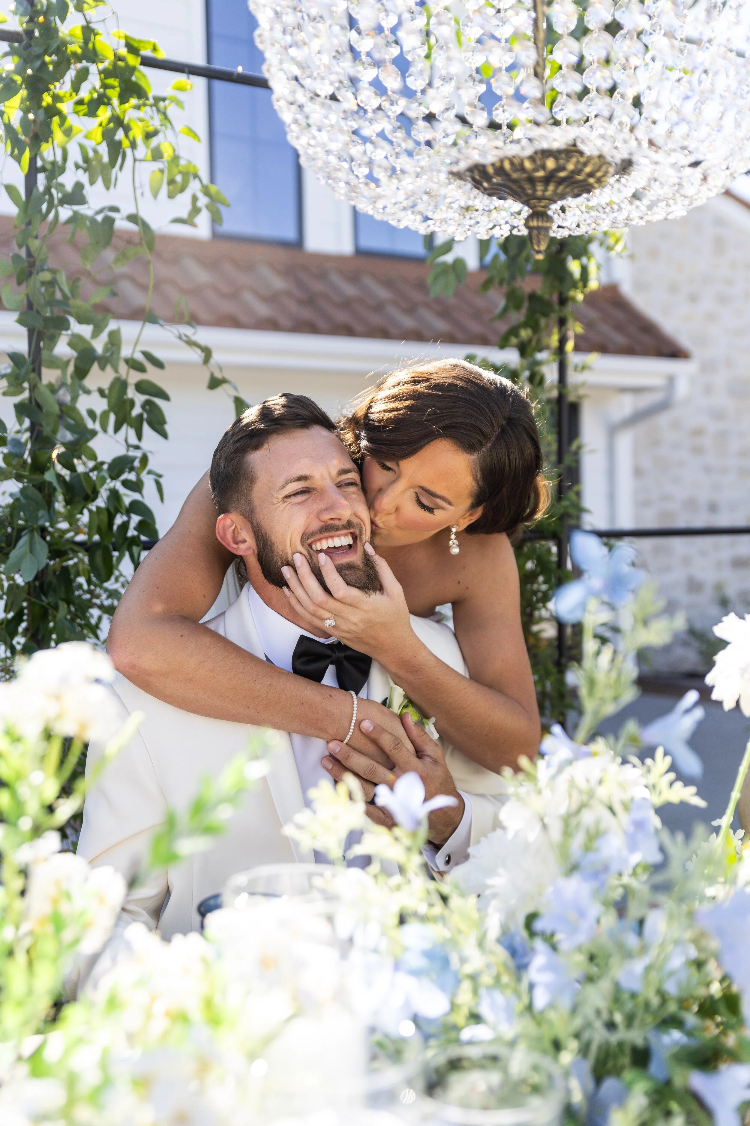 A bride kissing a groom on the cheek at a wedding reception with flowers and a chandelier overhead.