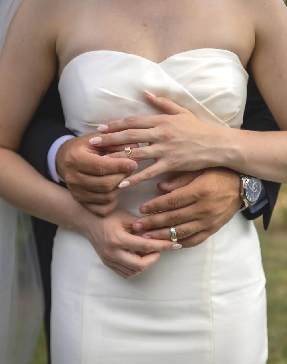 A bride and groom holding hands, with their hands placed on the bride's chest. They are wearing wedding rings and the bride is in a white wedding dress at their International wedding captured by Keri-Ann Gordon