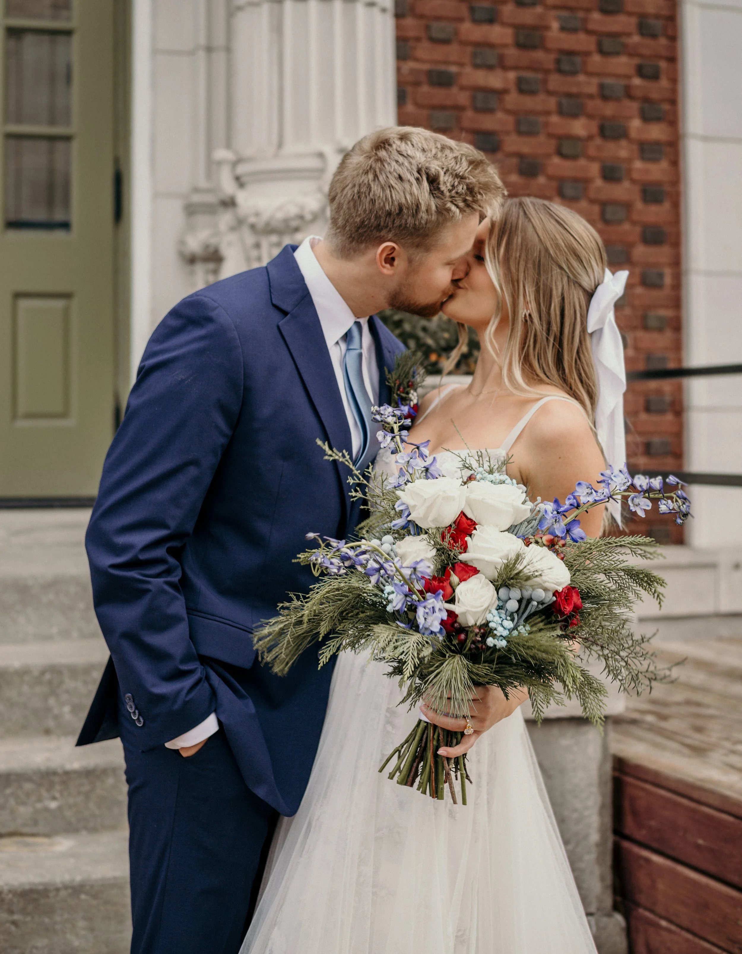 A newlywed couple sharing a kiss outdoors, the bride holding a large bouquet of flowers with white, red, and purple blooms and greenery, the groom in a navy blue suit, in front of brick and stone building steps. Kansas wedding photographer