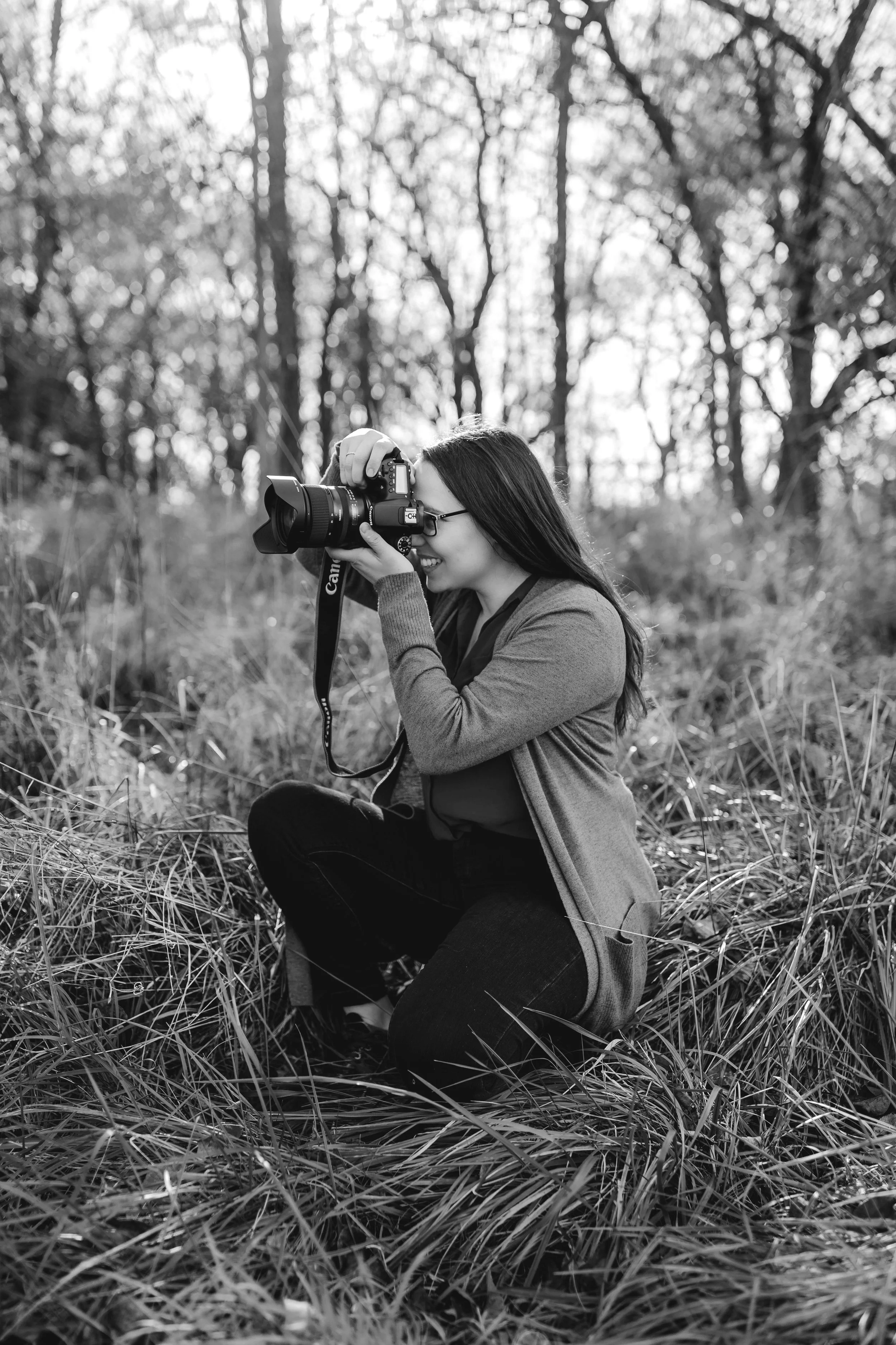 Kansas City Missouri wedding photographer KA Gordon kneels in tall grass, taking a photograph in a forested area with trees in the background, using a Canon camera.