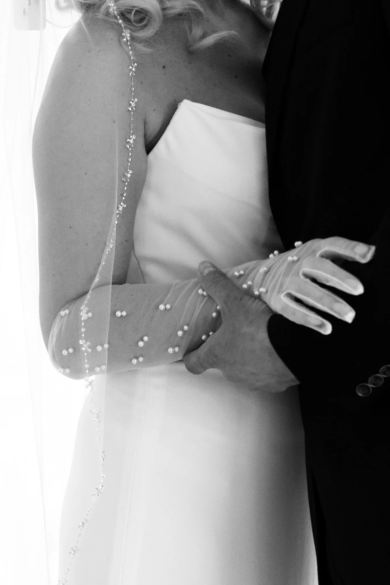 Close-up of a bride and groom holding hands during a wedding, with the bride wearing a pearl-embellished veil and gloves, and the groom in a dark suit, in black and white. Kansas and Kansas City wedding photography fine art and luxury