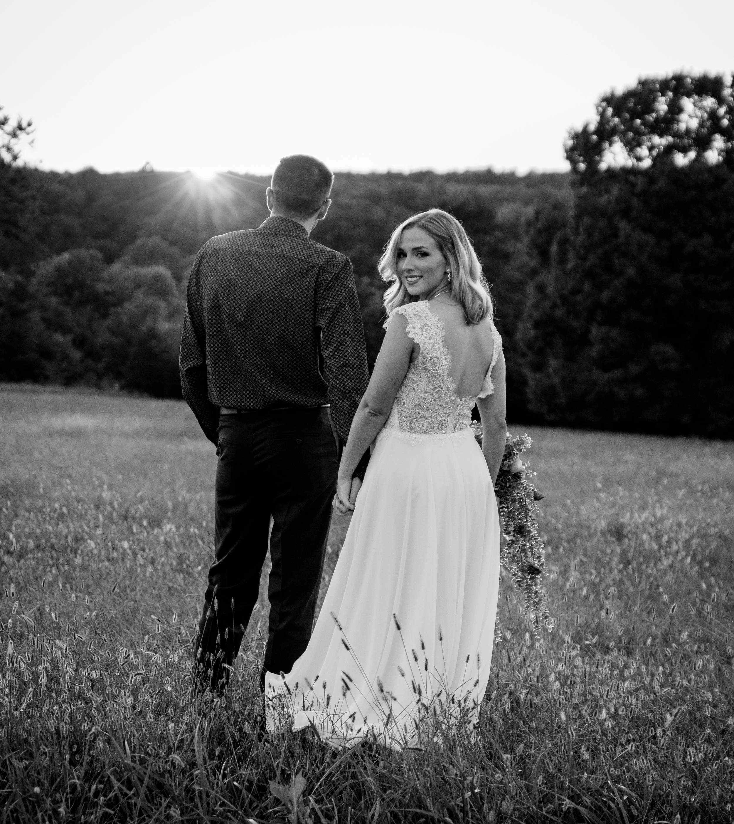 A bride and groom standing in a field at sunset, holding hands and looking back at the camera, with trees in the background.  Fine art wedding photographer in Kansas City and internationally.