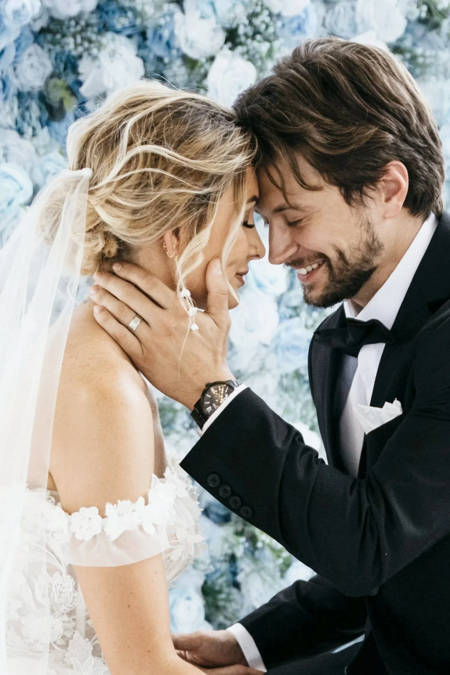 A bride and groom share a close, tender moment during their wedding, with their foreheads touching and eyes closed, against a backdrop of white and blue flowers. Kansas City luxury wedding photographer
