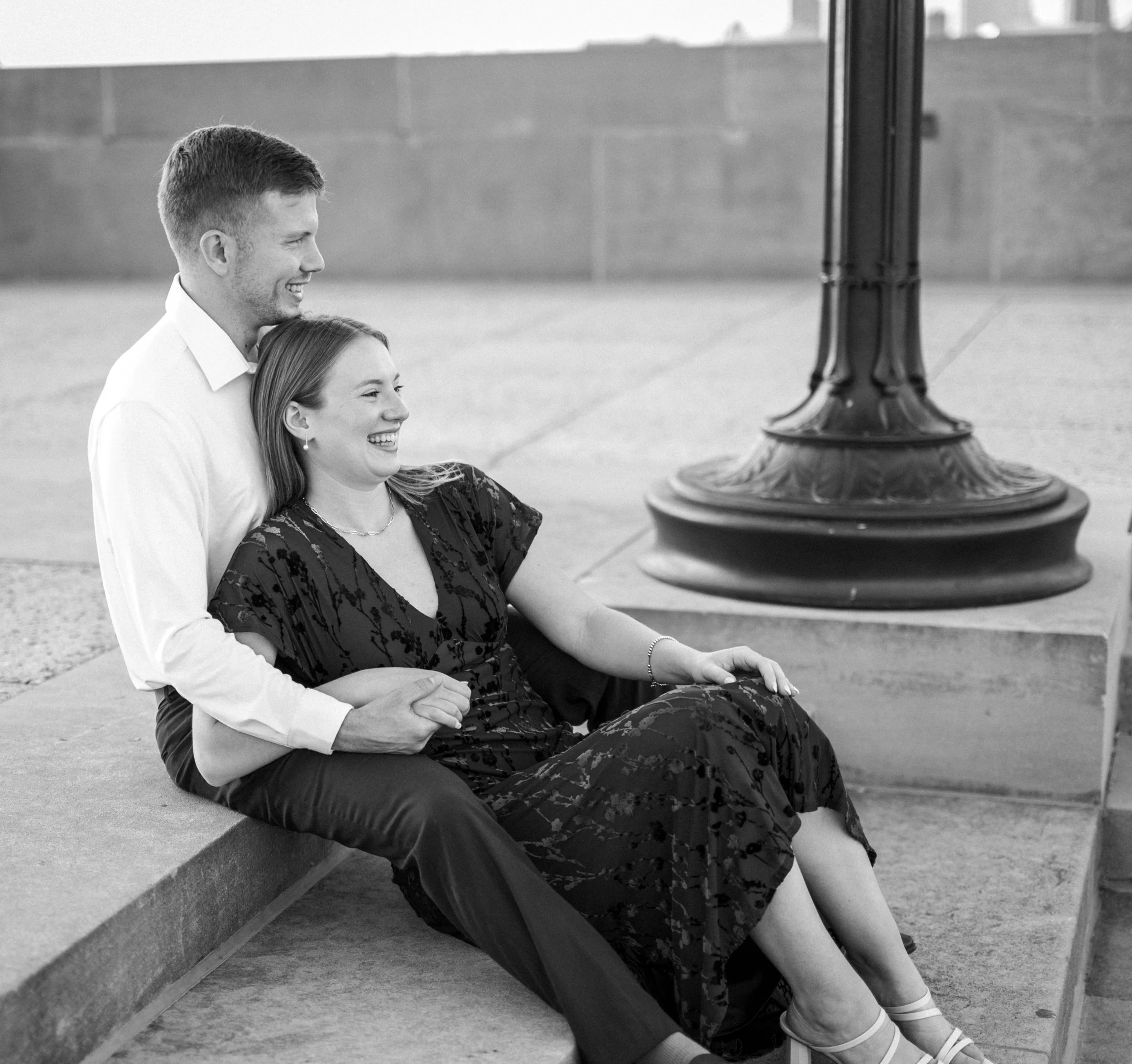 A happy couple sitting on a concrete ledge near a lamp post, smiling and laughing, black and white photo. Kansas and destination engagement photography