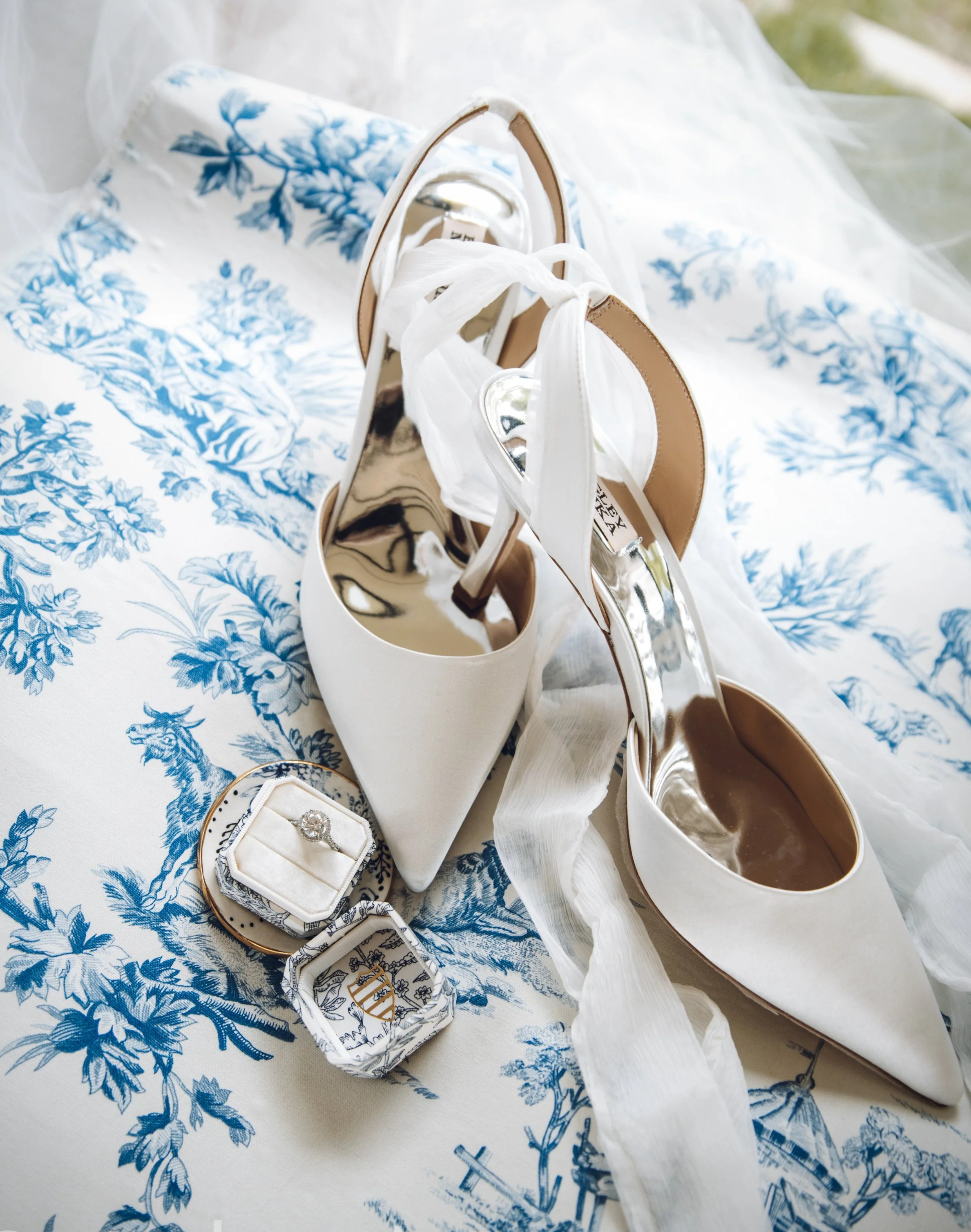 A pair of white pointed-toe high heels with ankle straps and ribbons, gold soles, resting on a blue and white floral patterned cloth, with a jewelry box containing a diamond ring nearby. Kansas city and international luxury wedding photographer