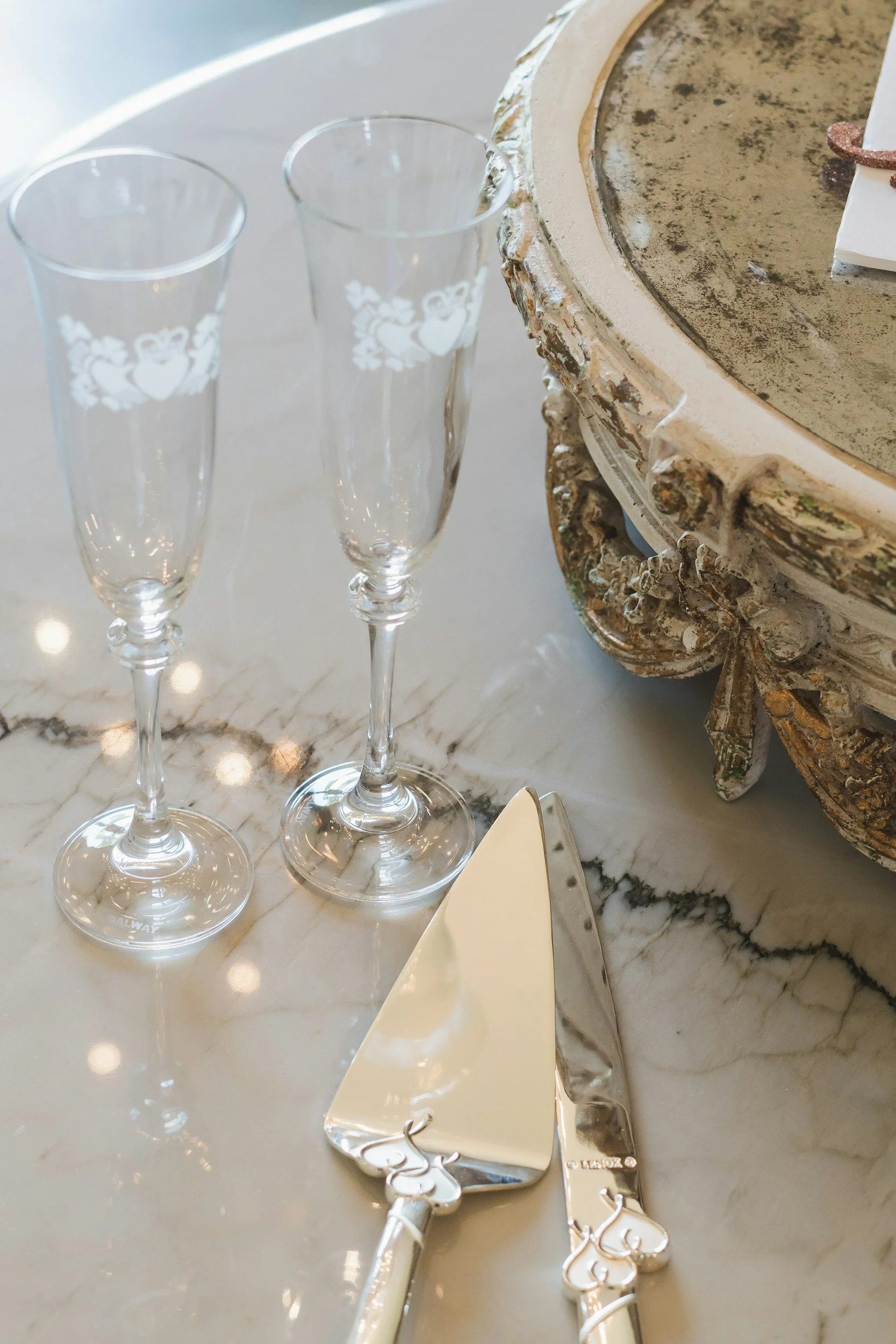 Two empty champagne flutes with decorative white heart and floral patterns, a wedding cake server and a knife with ornate handles, and a decorative ornate mirror or frame on a white marble table. Kansas editorial wedding photographer