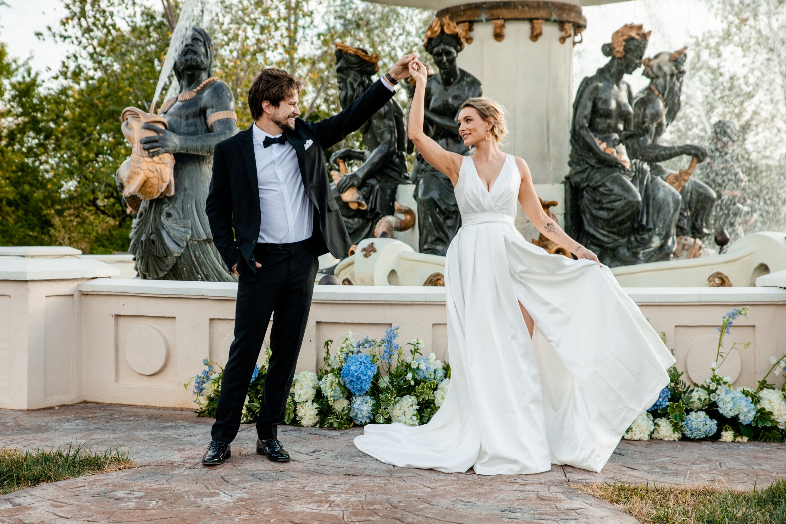 A couple in wedding attire dancing together outdoors in front of a decorative fountain with bronze statues, and blue and white flowers at the base of the fountain. European Luxury wedding and elopement photographer