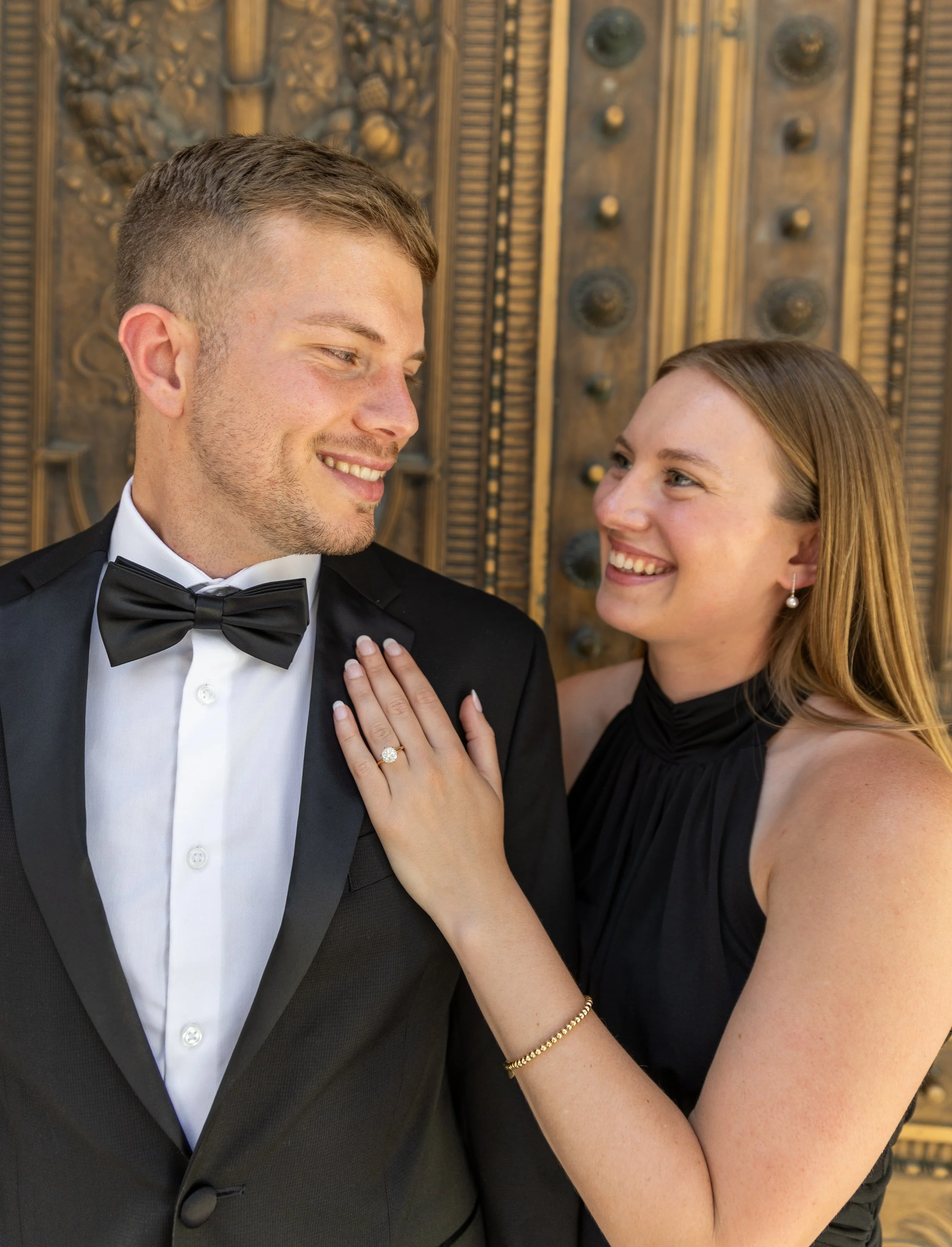 A couple dressed in formal attire, with the woman showing her engagement ring, standing in front of a decorative wooden door. Kansas city engagement and wedding photographer