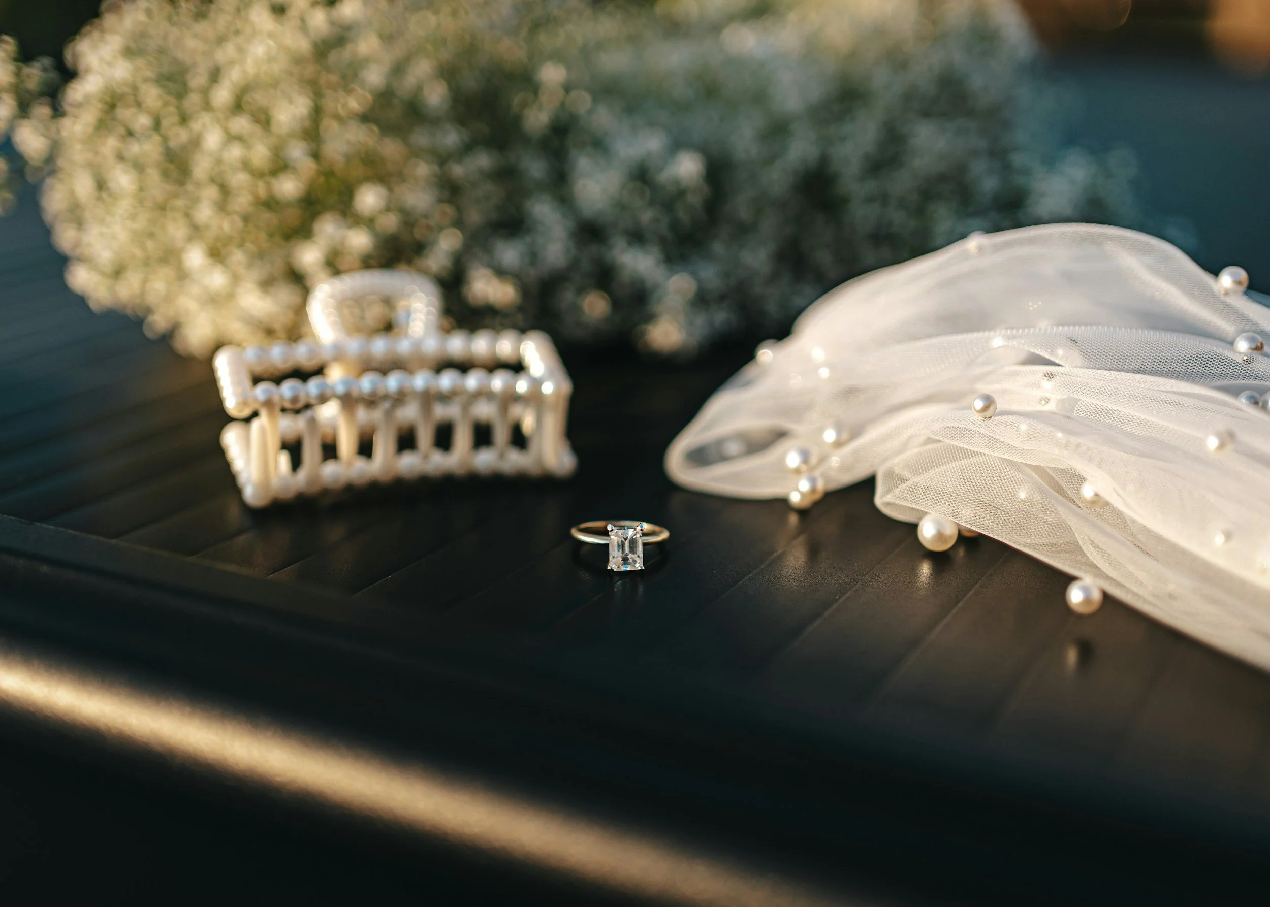 Close-up of a diamond ring, pearl bracelet, and a white veil with pearls, arranged on a dark surface, with a blurred background of flowers. Kansas City elopement and intimate wedding photographer