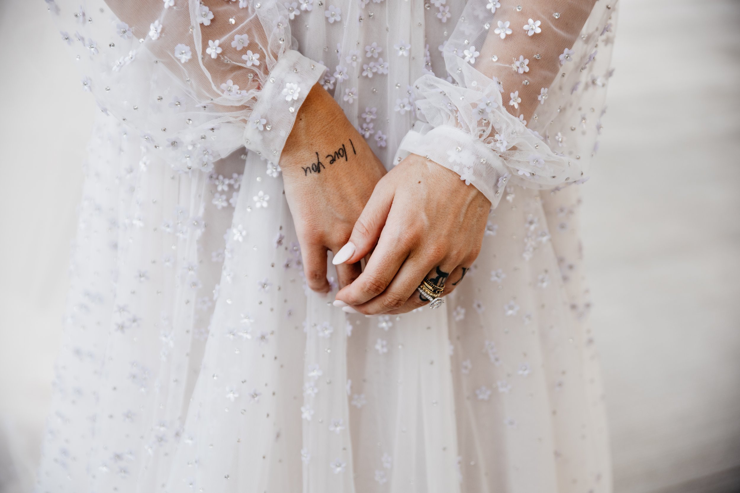 Close-up of a person wearing a white, sheer dress with floral embroidery, their hands clasped together, displaying rings and a tattoo that reads "Love you" on their wrist. Kansas City luxury wedding photographer