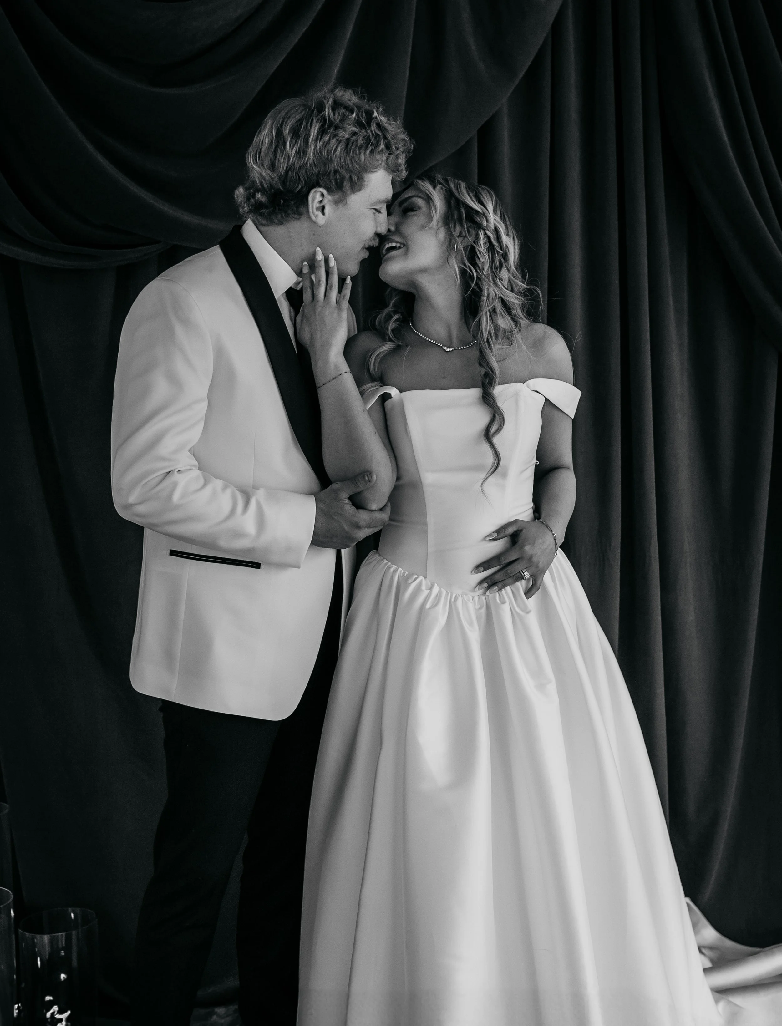 A black and white photo of a bride and groom in wedding attire standing close together, with their noses touching and smiling at each other, in front of dark curtains. Fine art wedding photography in Kansas and worldwide