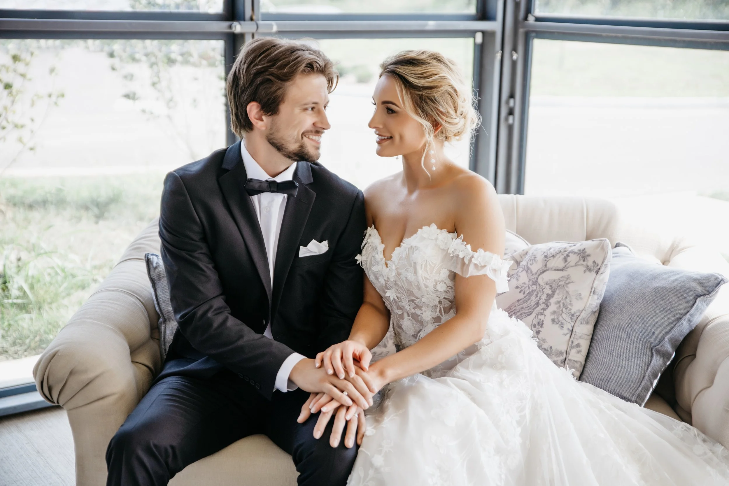 A bride and groom sitting on a beige sofa, holding hands and smiling at each other, with large windows behind them showing a green outdoor scene. editorial wedding photography in Kansas