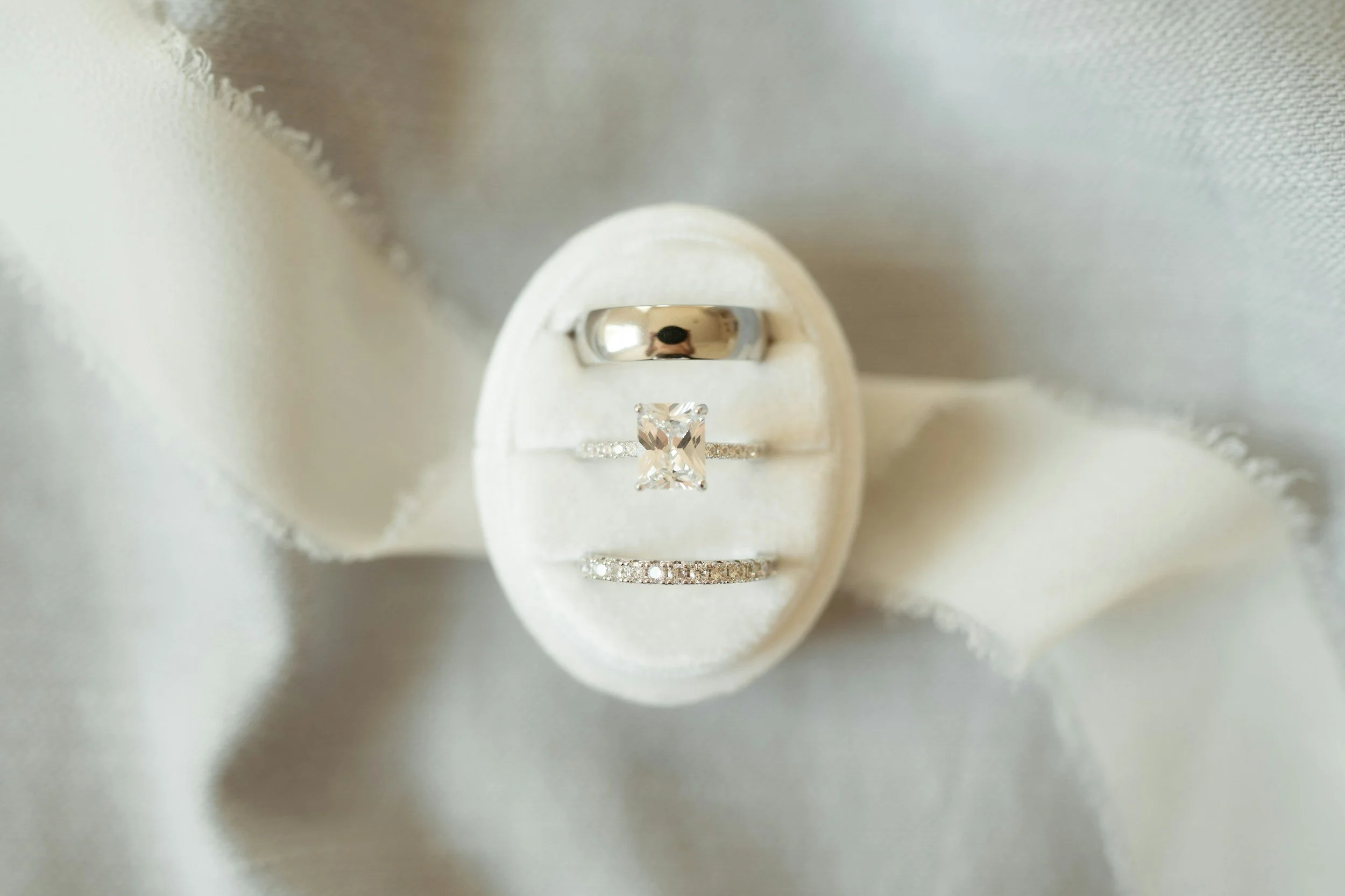 Close-up of three rings: a silver band, a solitaire diamond ring with a square cut stone, and a diamond-studded band, placed on a white fabric background. Kansas City Missouri and destination wedding photography