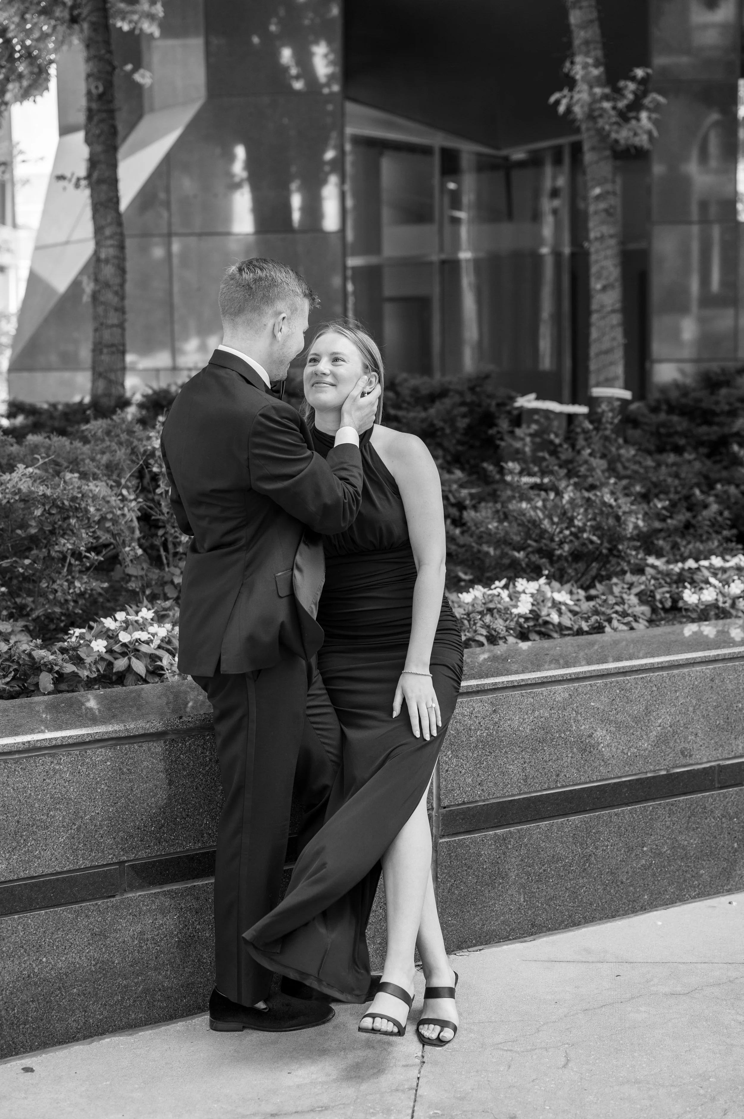 A man in a suit gently holding a woman's face with both hands, as they share a tender moment outdoors near a building and garden. The woman is wearing a long, elegant dress with a slit and sandals, and they are standing against a concrete planter fil